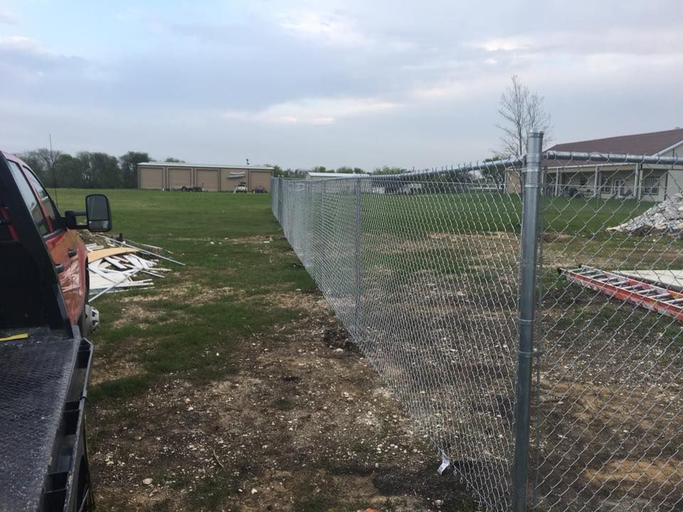 Chain-link fence along a dirt path beside a grassy field and buildings under a cloudy sky