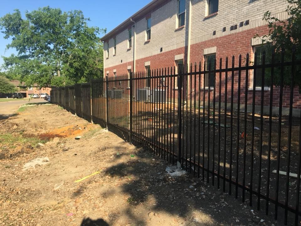 Black metal fence beside a beige and brick apartment building along a dirt path in bright sunlight