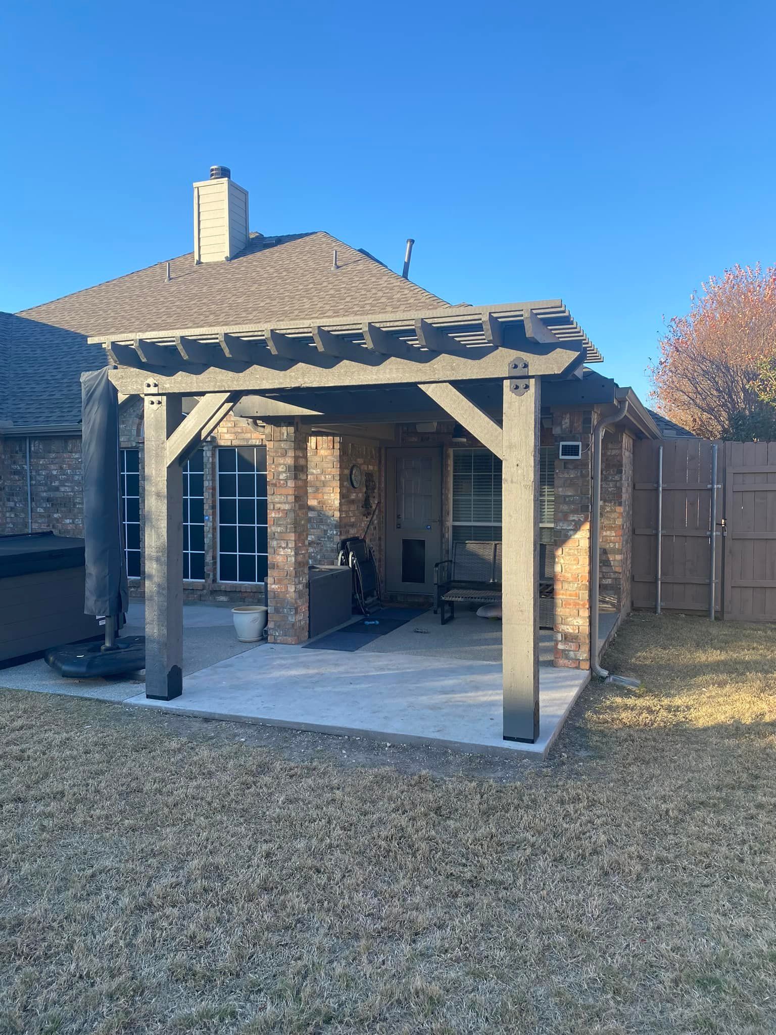 Backyard covered patio with wooden posts, stone fireplace, and shingle roof under a clear blue sky