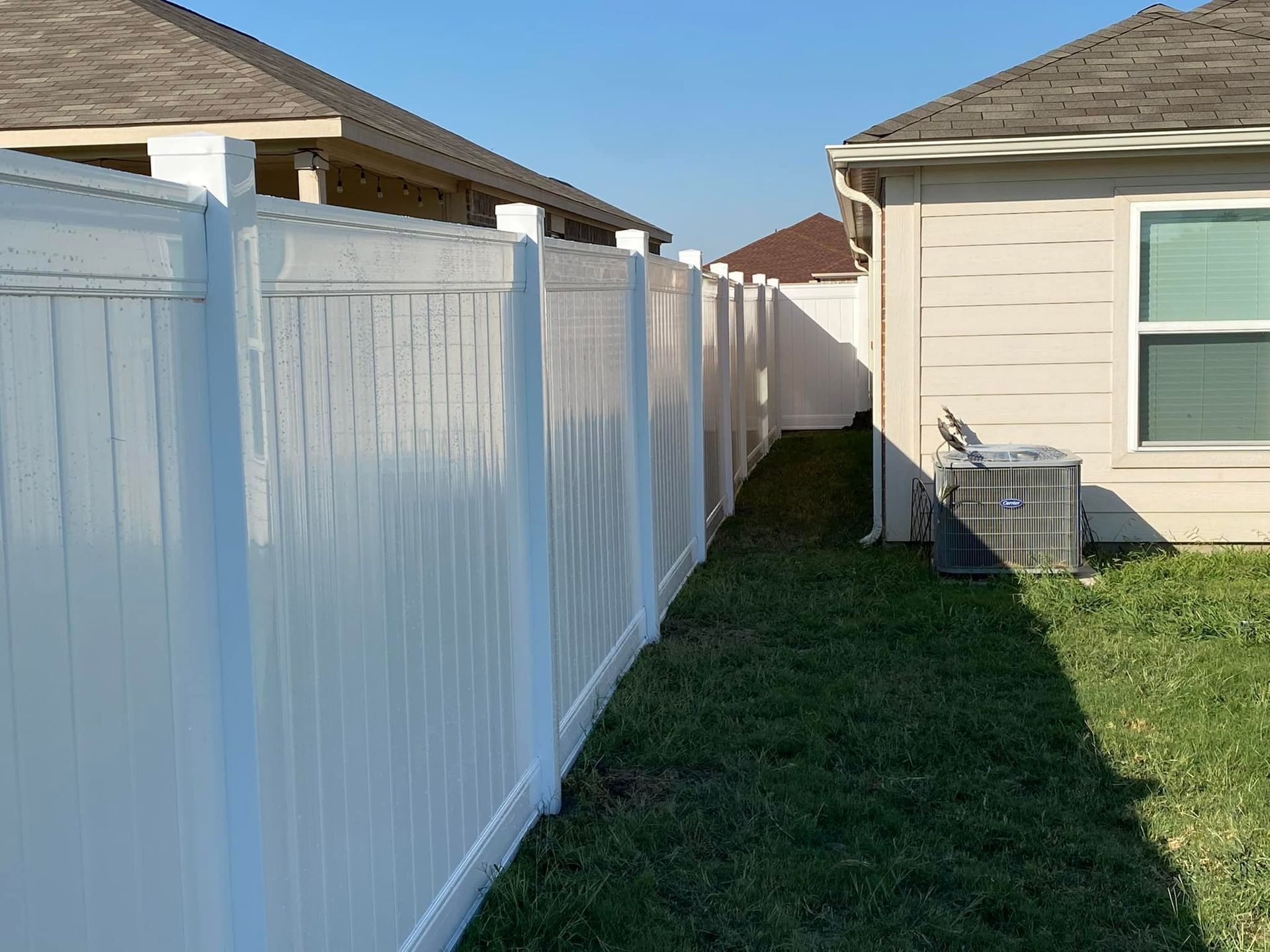 Narrow grassy side yard between a white vinyl fence and a beige house on a sunny day