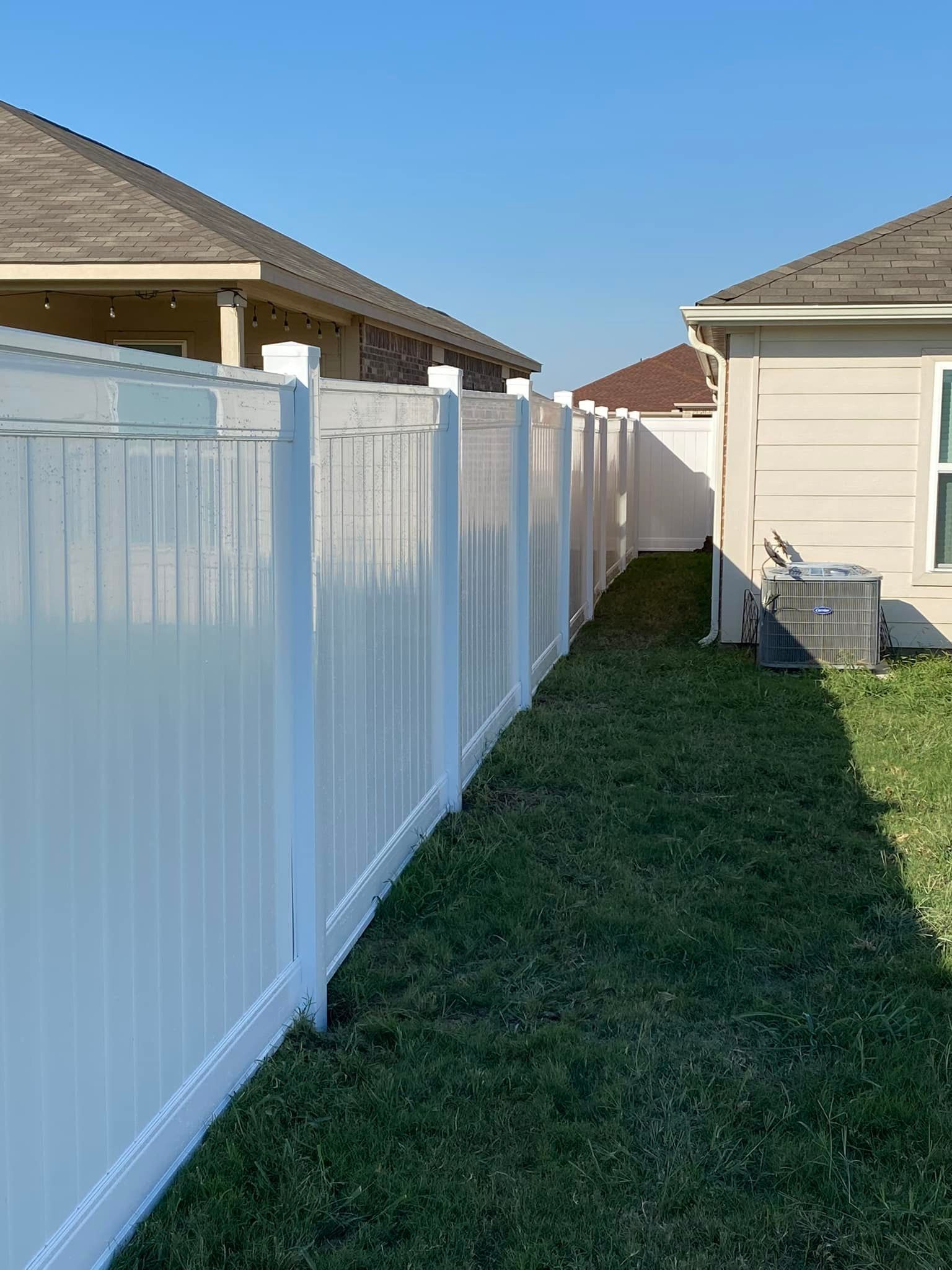Narrow grass strip between white vinyl fence and beige house under a blue sky.