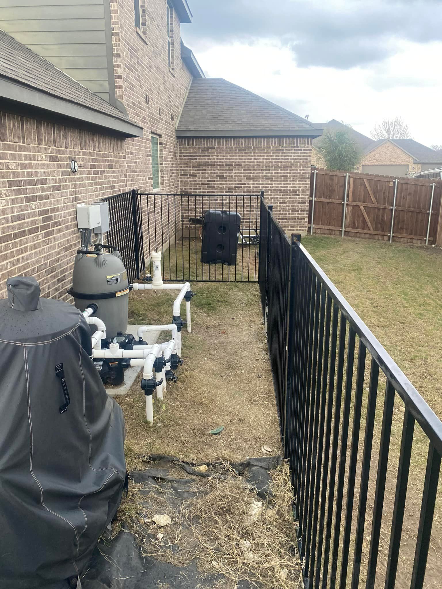 Brick house side yard with black metal fence, gravel path, patio grill, and wooden gate in the background