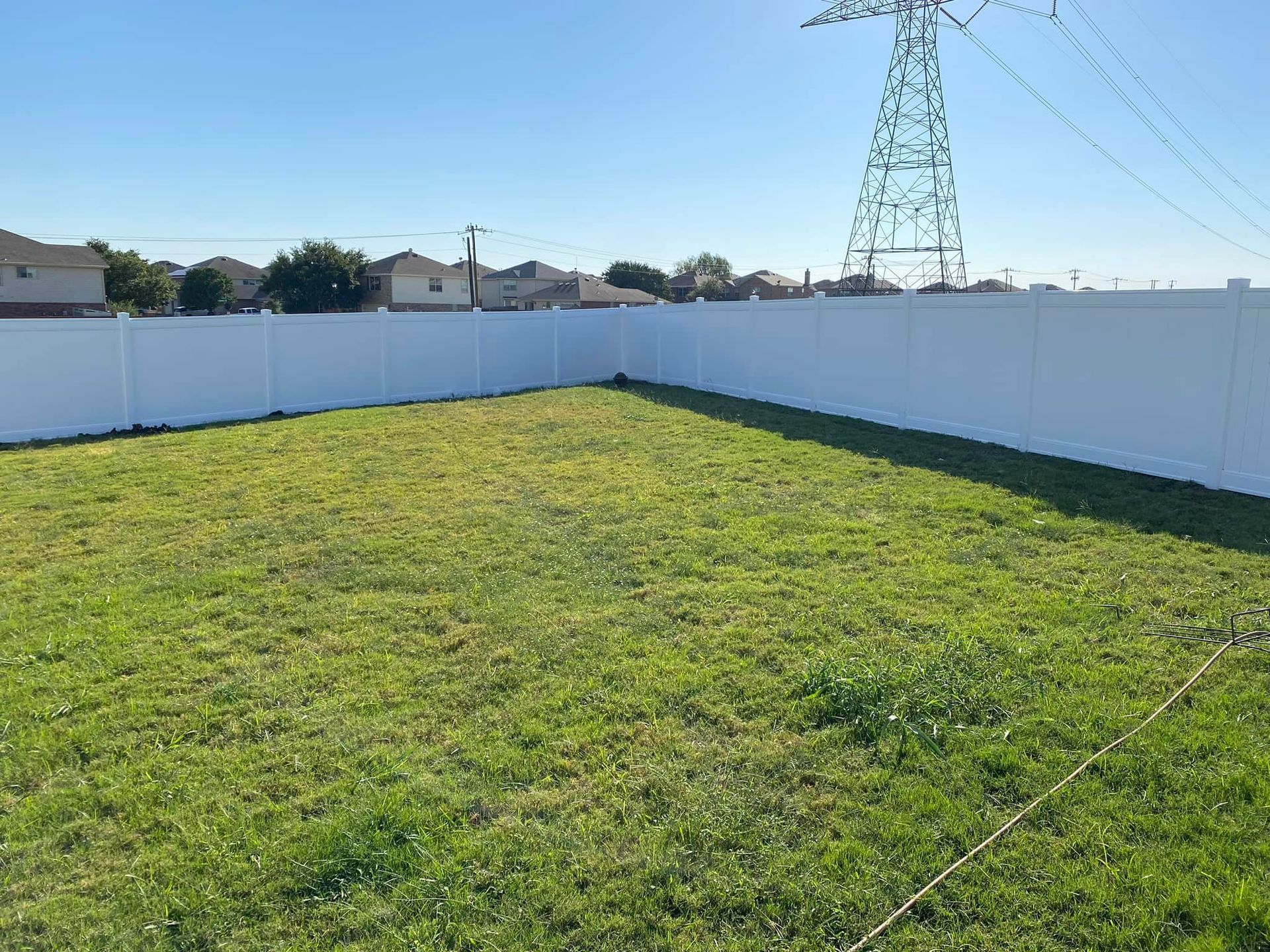 Empty grassy backyard with white vinyl fencing and power lines overhead under a clear blue sky