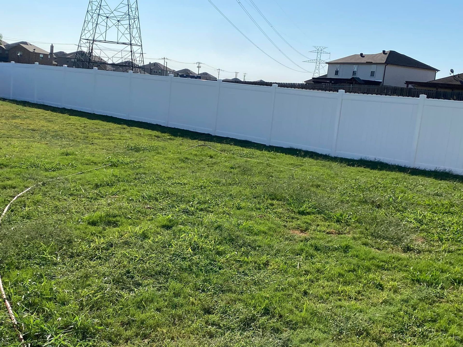 Grassy field beside a white fence under a blue sky, with power lines and houses in the background