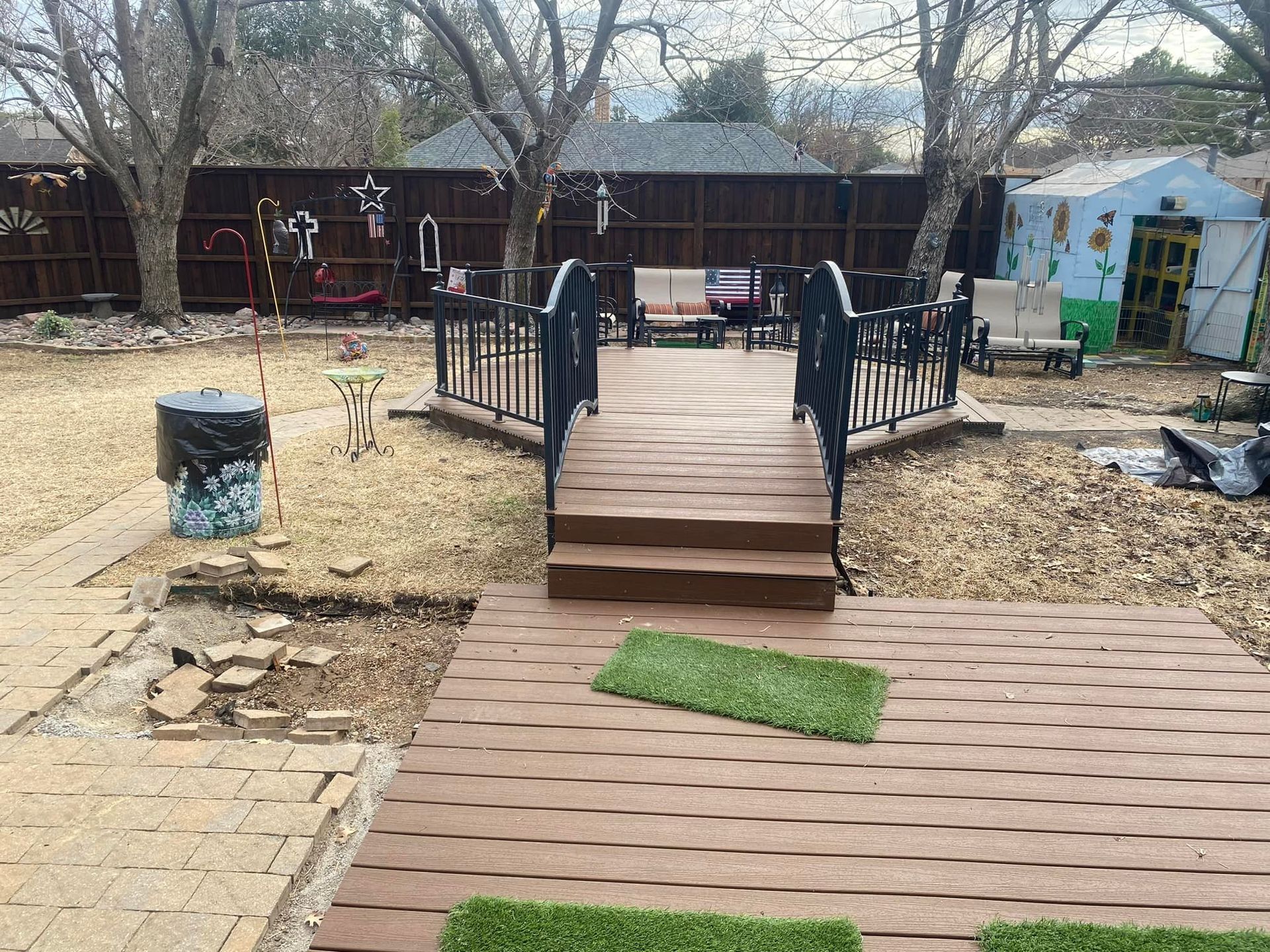 Backyard deck with wooden steps, black railings, and a small green mat on the patio facing a fenced yard.