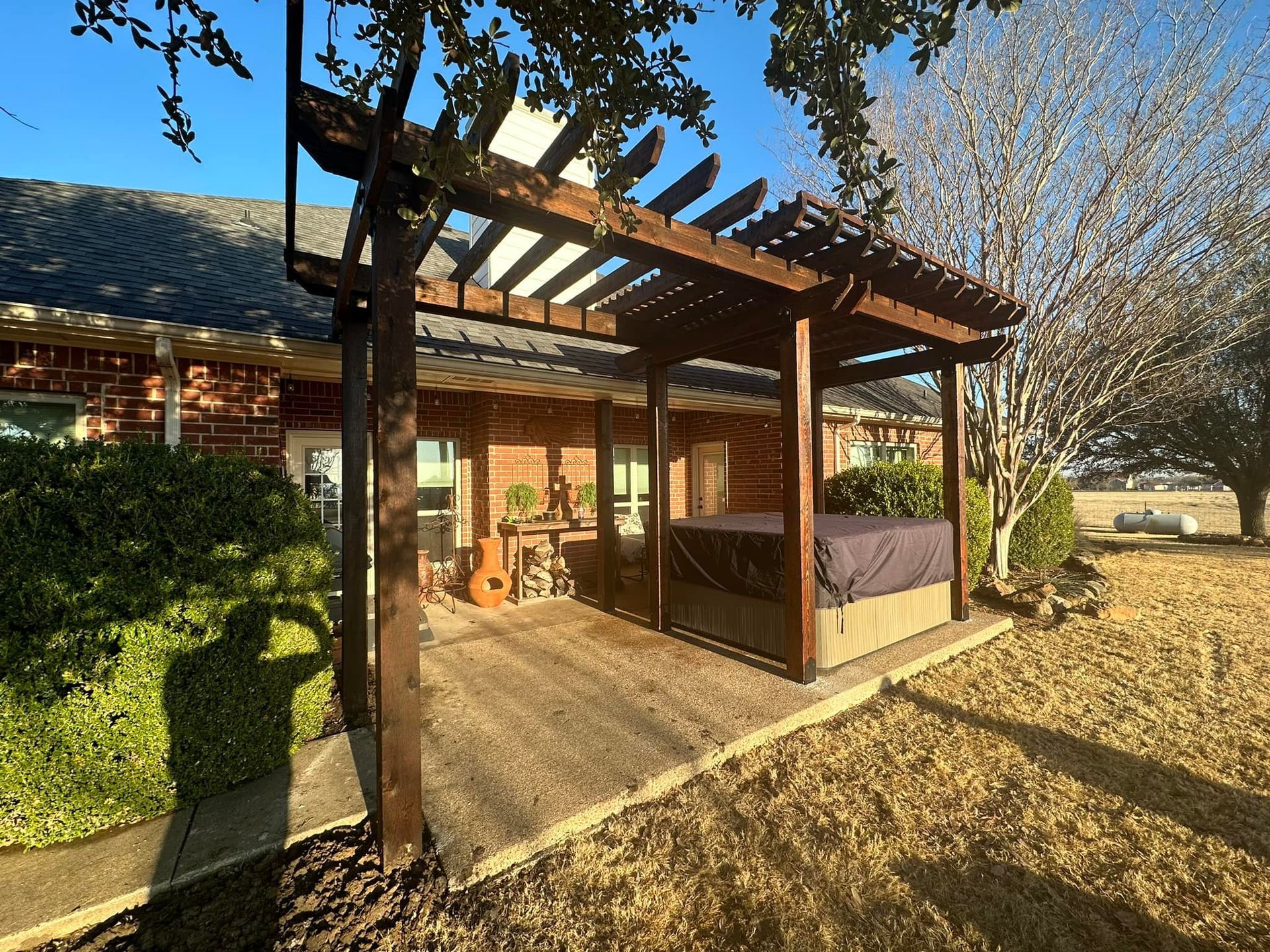 Wooden pergola on a sunlit patio beside a house, with a table and garden greenery.