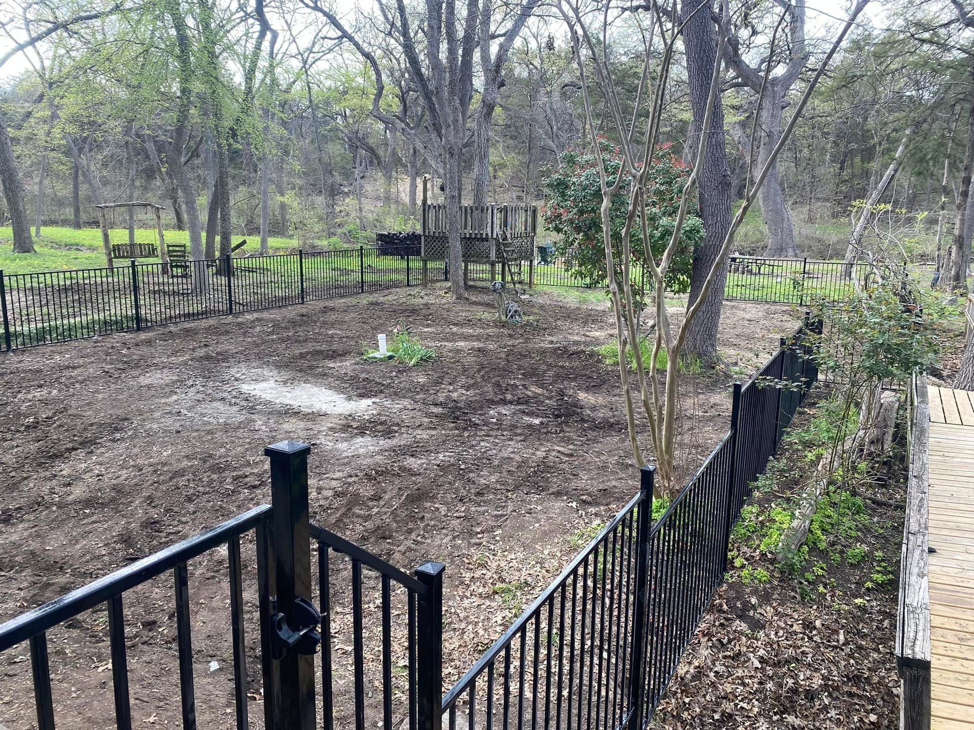 Black metal fence around a bare yard with trees and a small wooden structure in the background