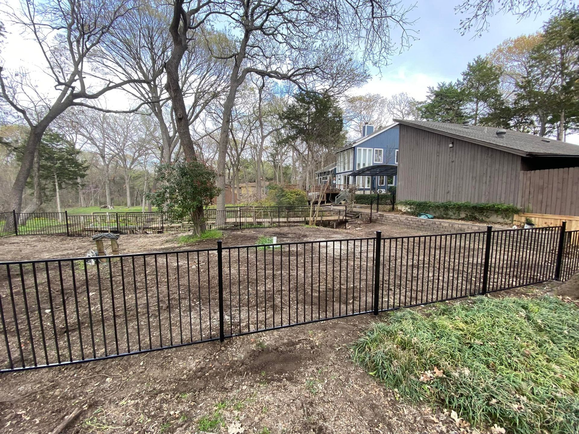 Black metal fence in a wooded yard with a barn-like shed and trees in the background