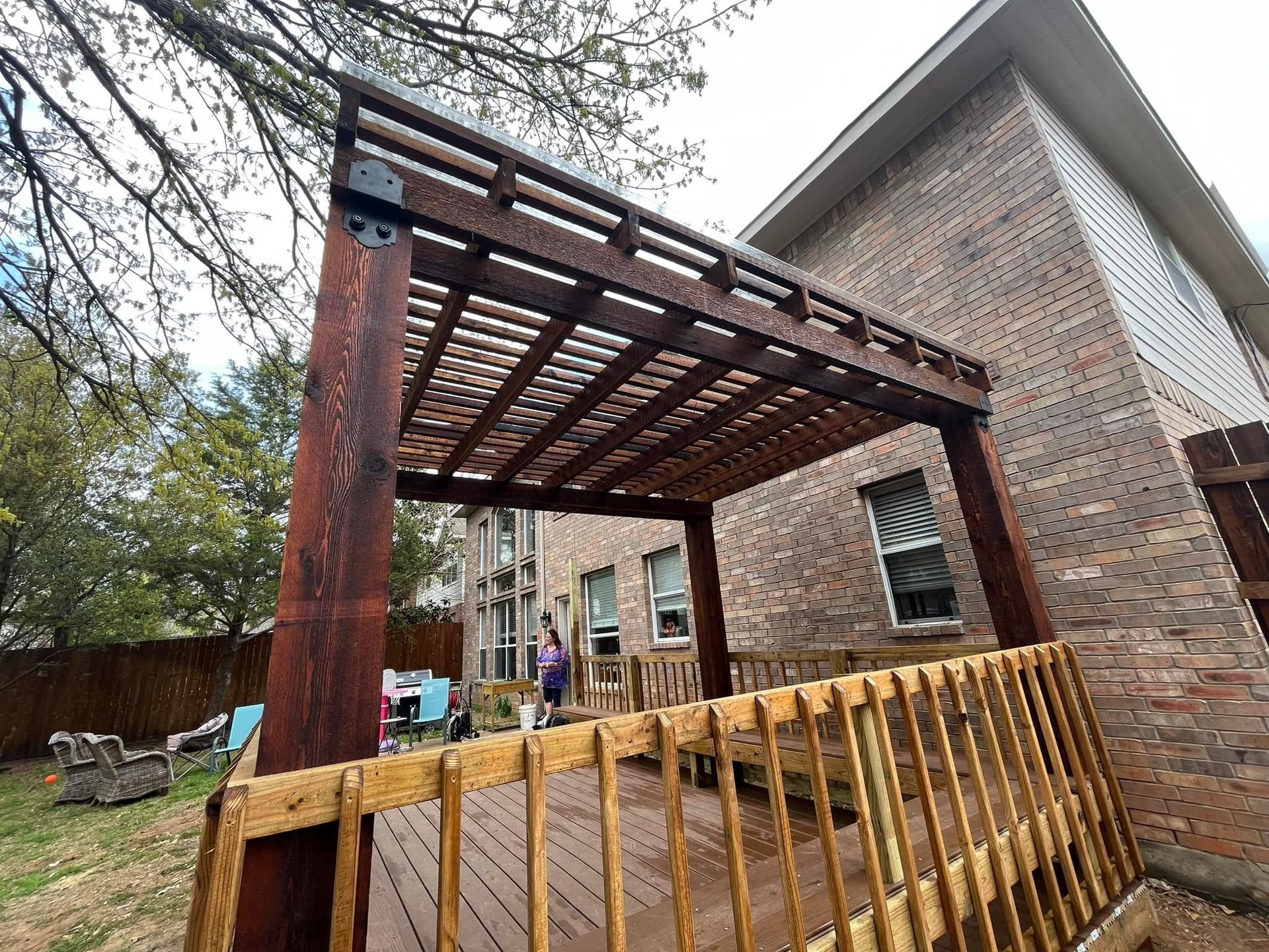 Wooden pergola frame beside a brick house, with a yellow barricade in front and a yard in the background.