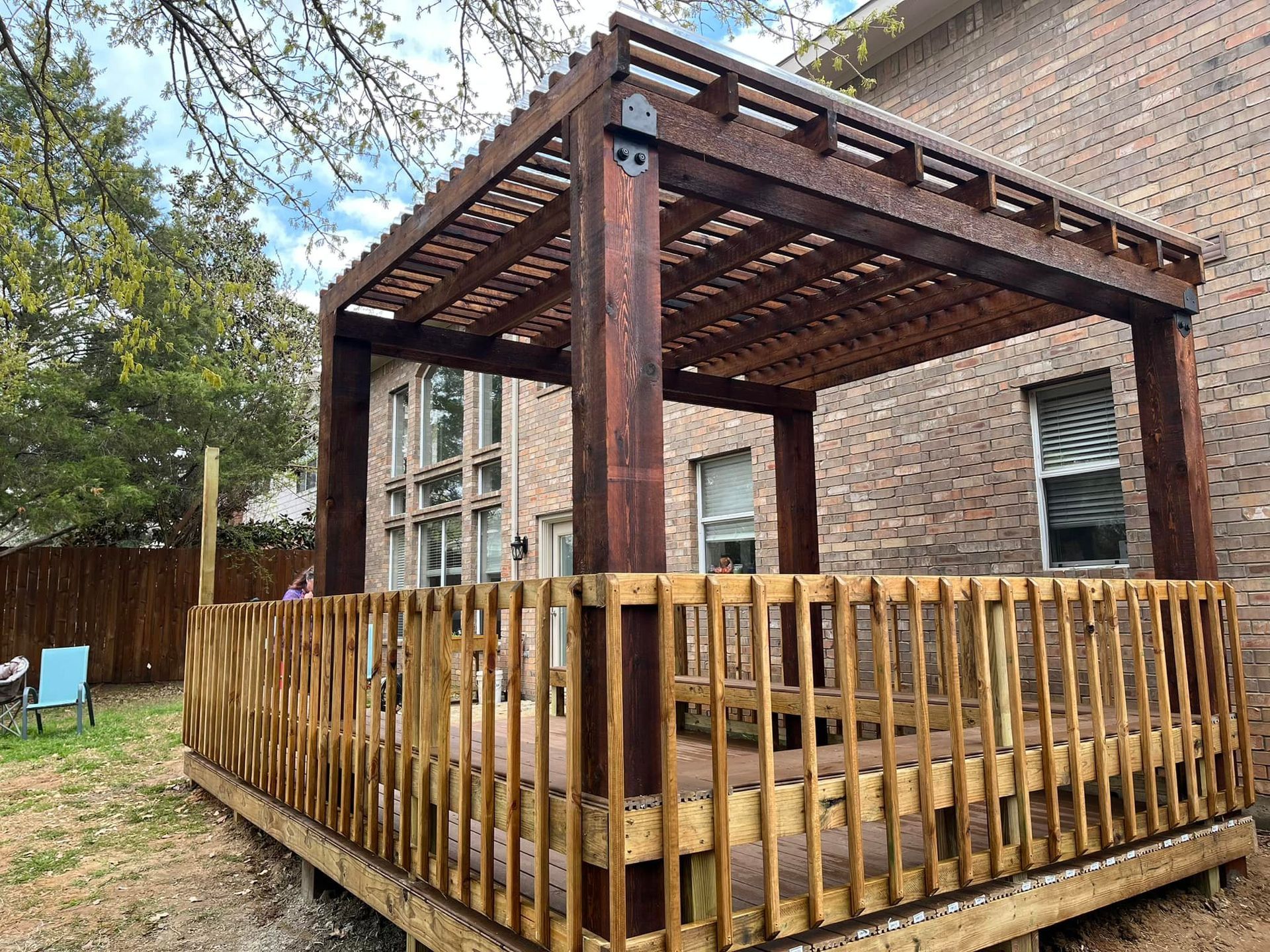 Wooden deck with pergola beside a brick house, surrounded by a fenced backyard and trees.