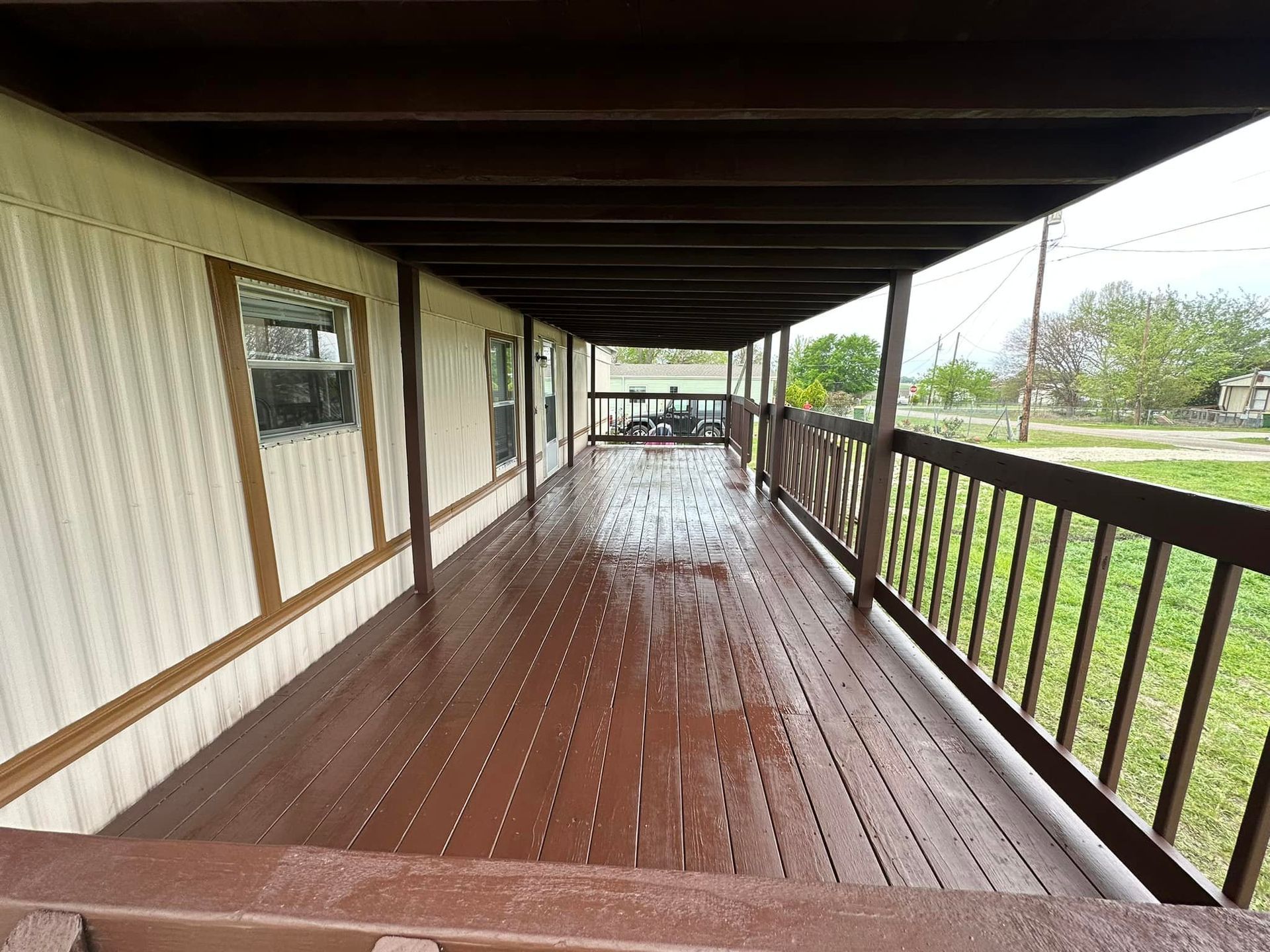 Long covered wooden porch with wet floor, railing, and grassy yard outside