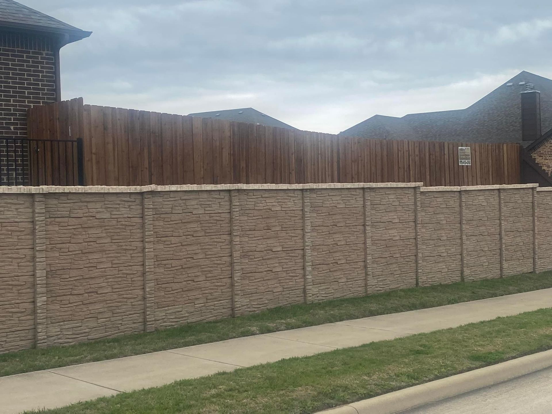 Residential backyard with tan brick wall, wooden fence, and cloudy sky above a sidewalk and grass strip