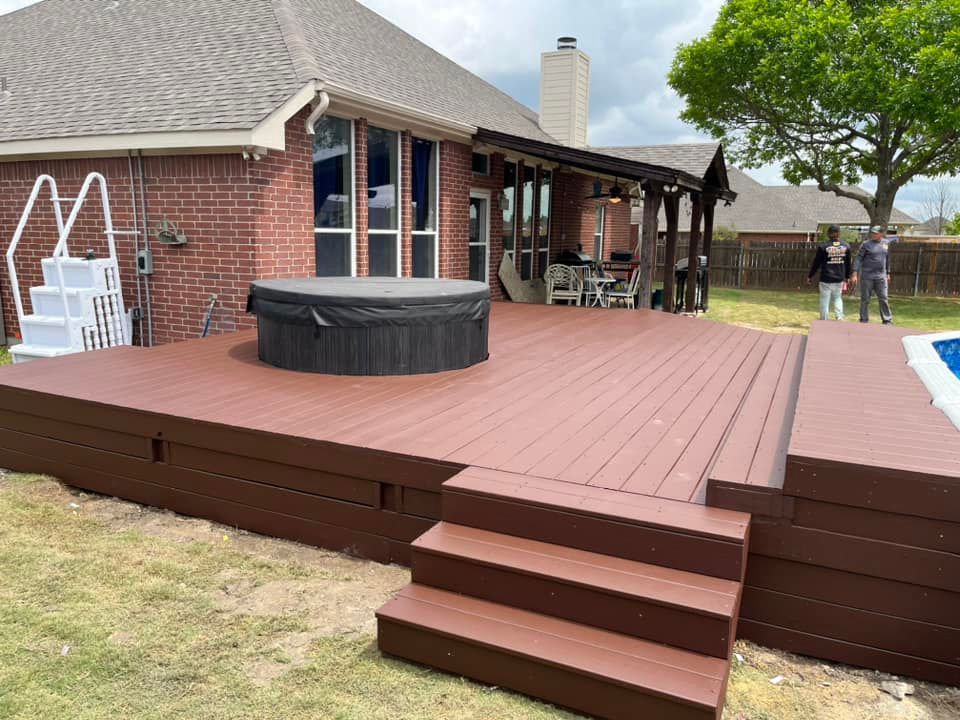 Backyard deck with steps, hot tub, covered patio, and brick house; two people stand near the fence.