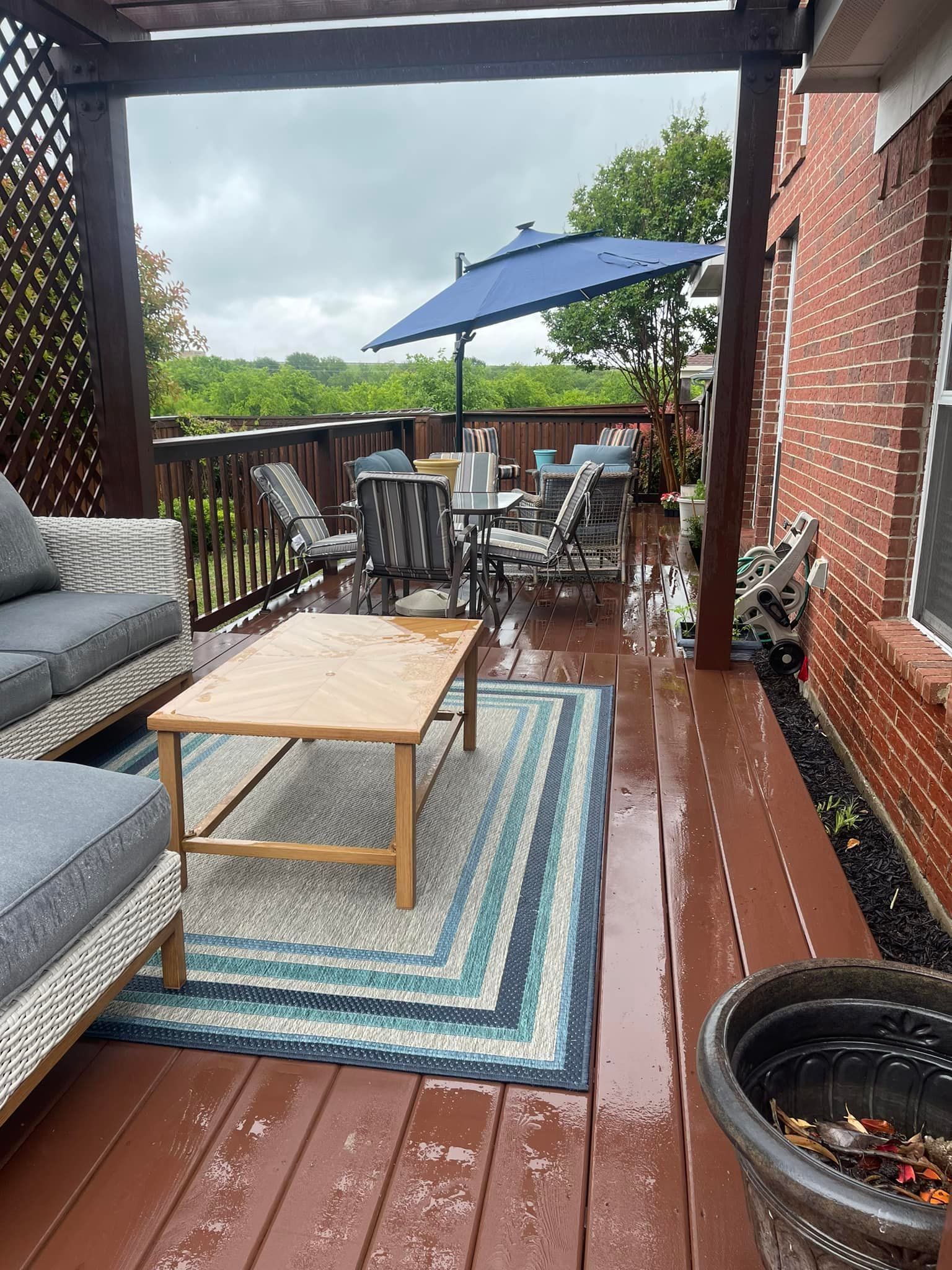 Covered patio with gray seating, wooden table, blue rug, and patio umbrella overlooking greenery