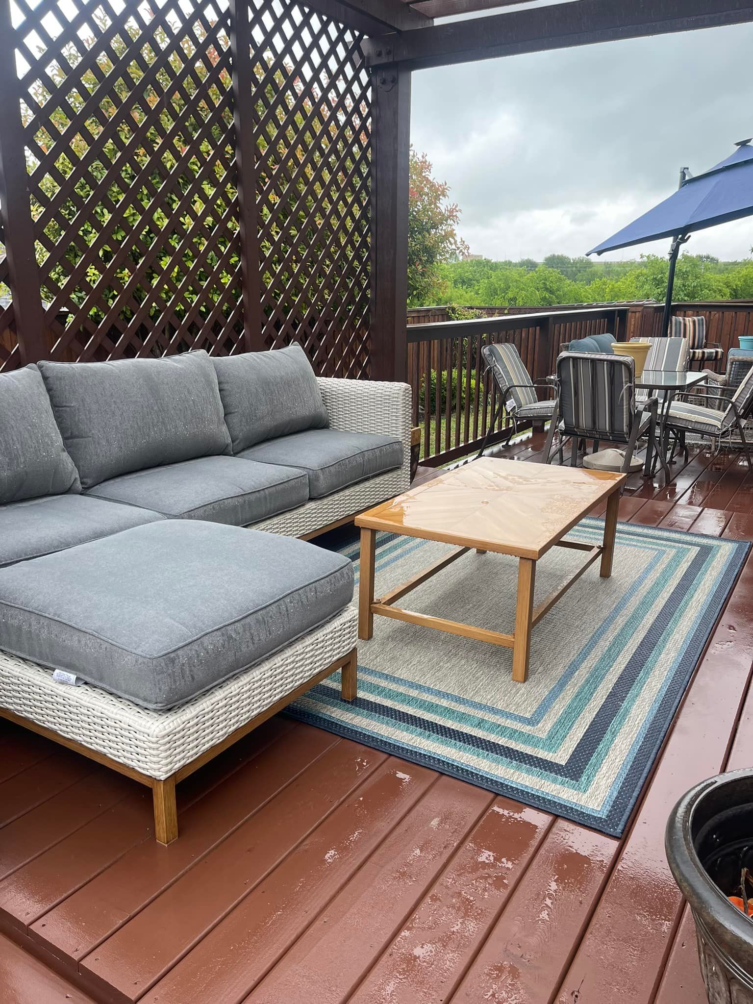 Patio with gray cushioned sofa, wooden coffee table, striped rug, and dining set on wet wooden deck