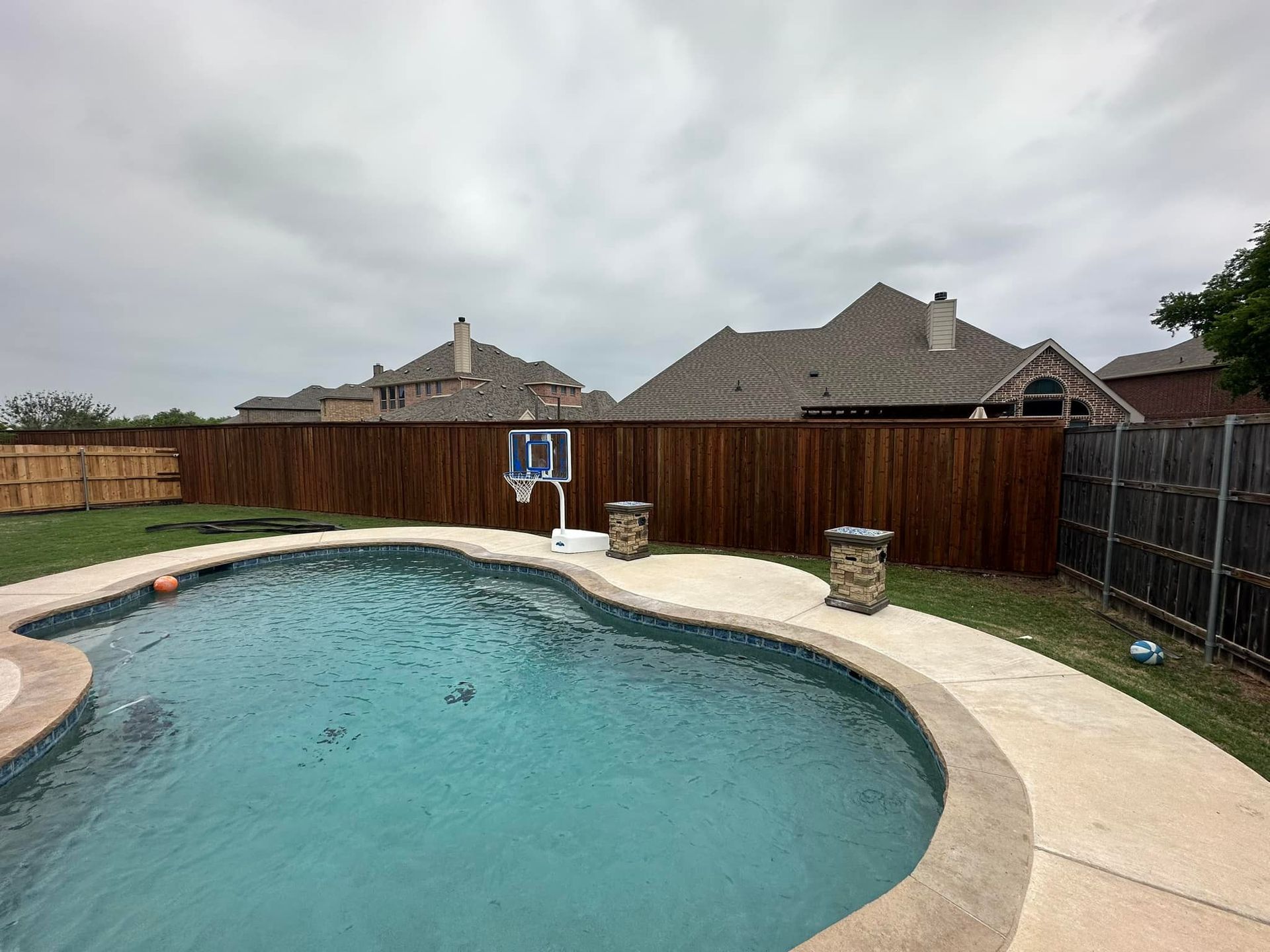 Backyard swimming pool with curved stone deck and wooden fence under a cloudy sky