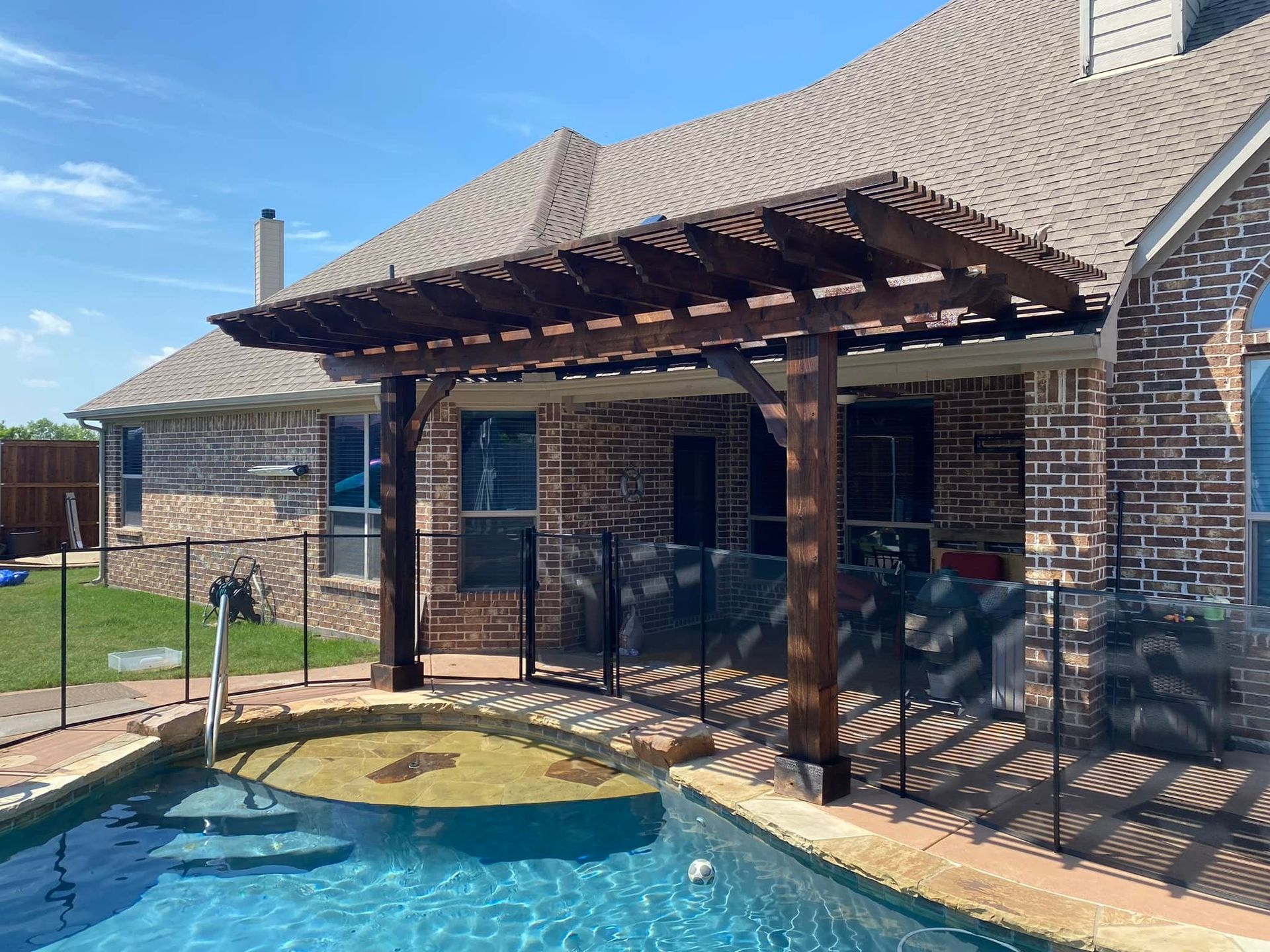 Backyard pool beside a brick house with a wooden pergola and covered patio.
