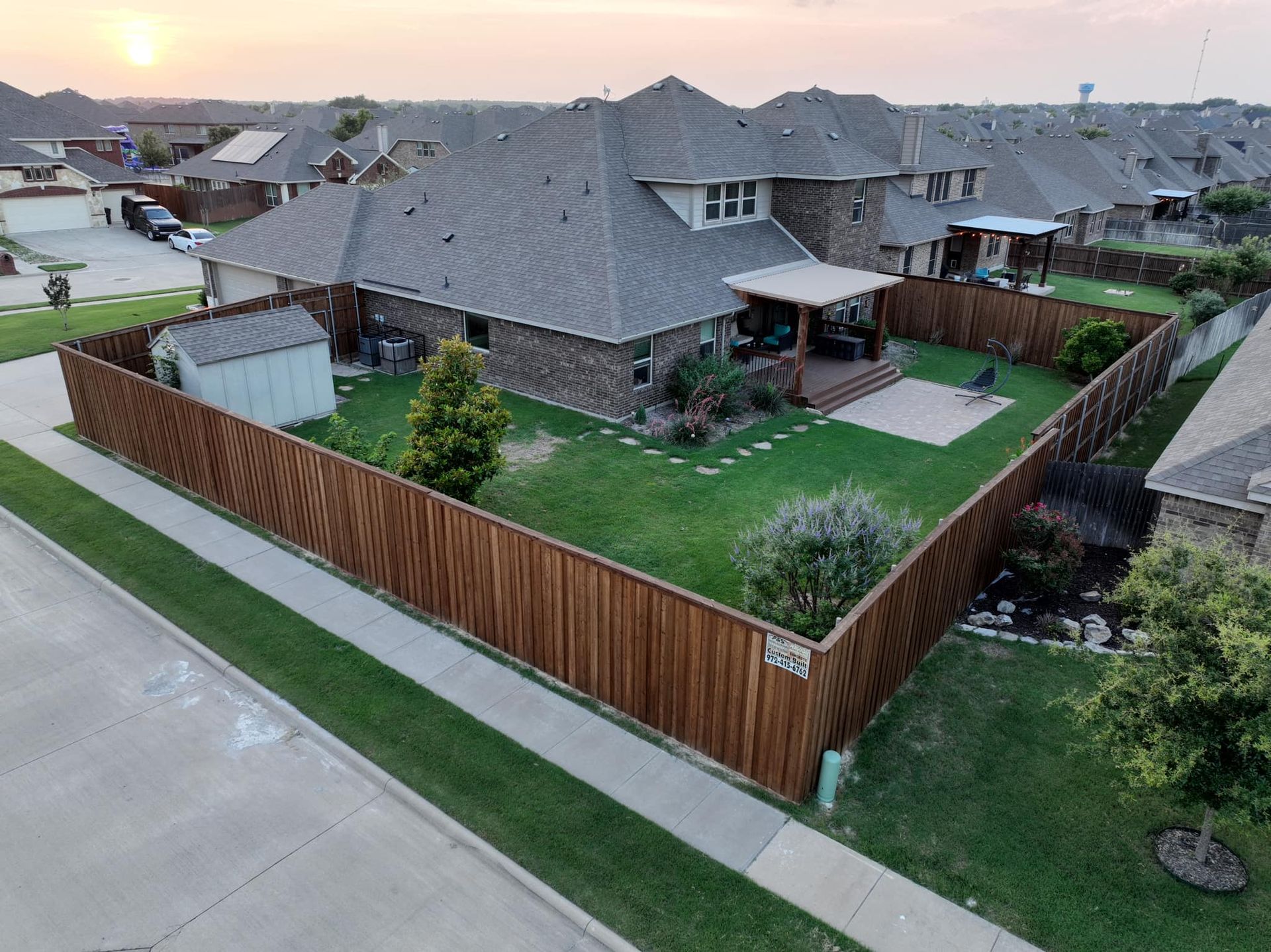 Suburban house with gray roof, fenced green yard, and driveway at sunset