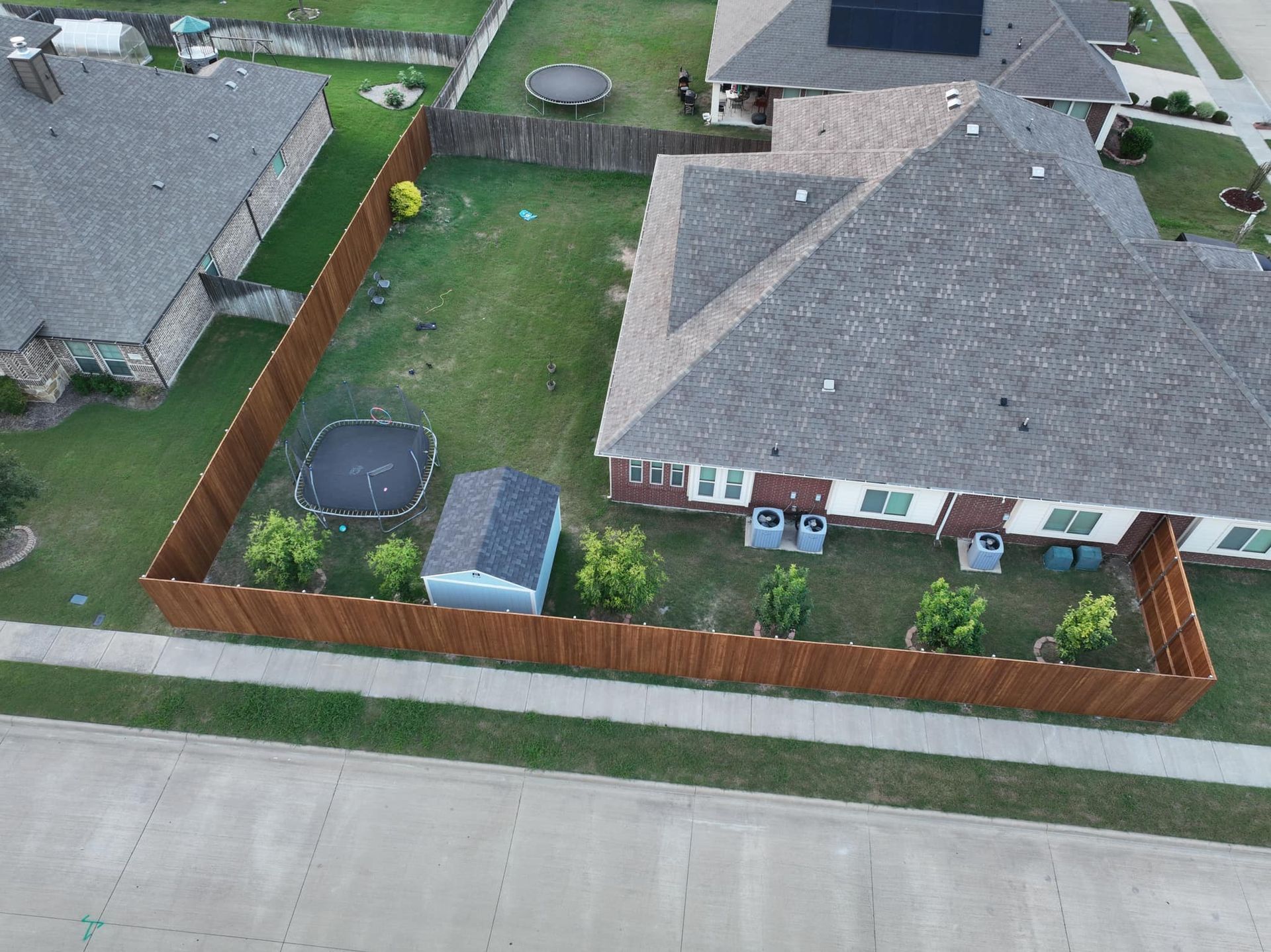 Aerial view of a suburban house with fenced backyard, driveway, and neighboring homes along the street.