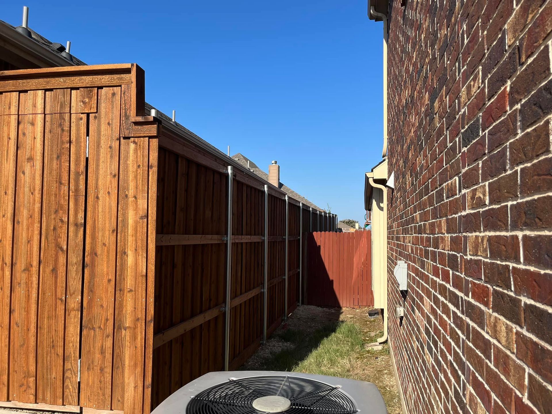 Narrow alley between a wooden fence and brick wall under a clear blue sky.