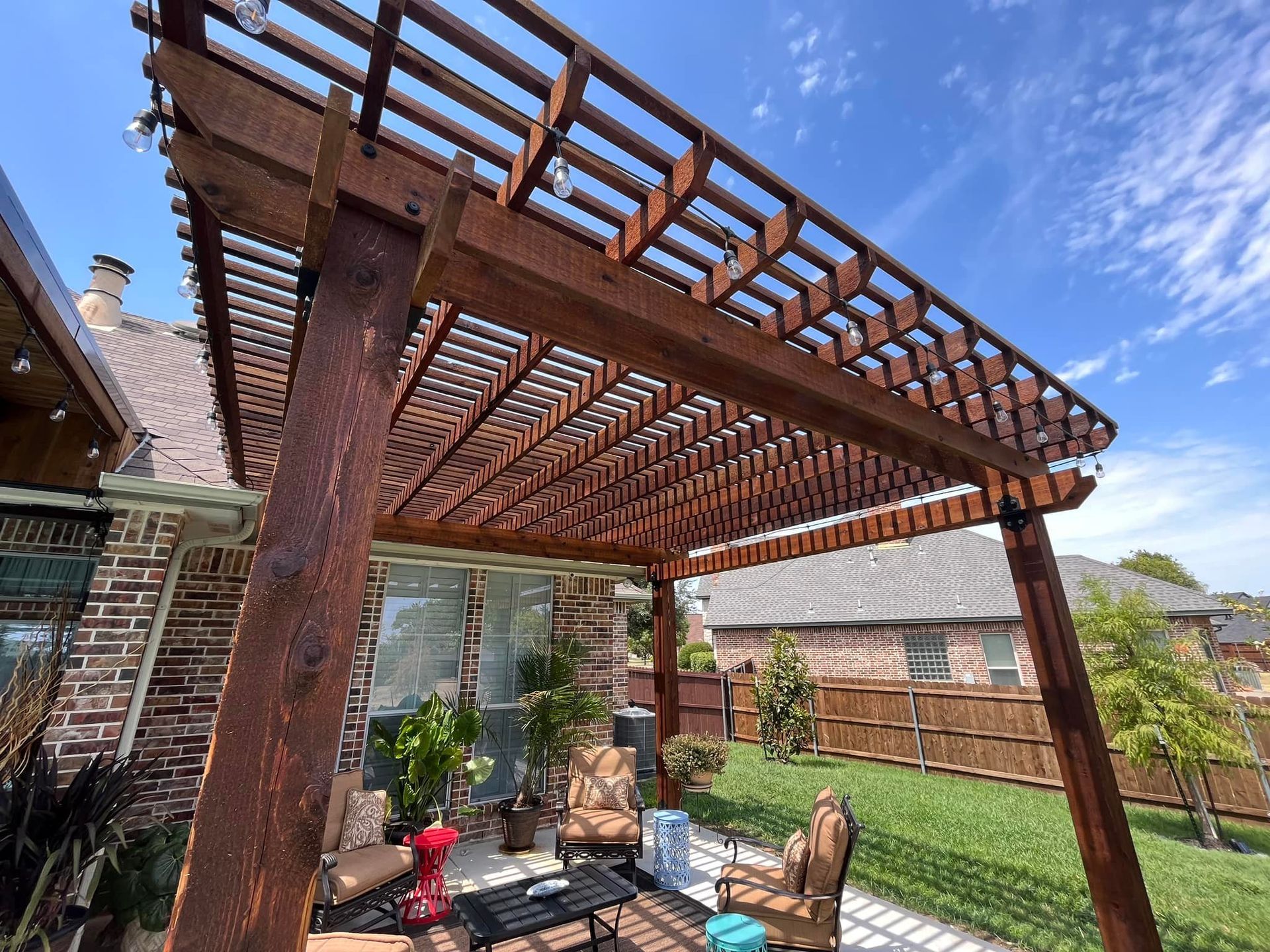 Wooden pergola over a brick patio with chairs, potted plants, and a lawn beside a house
