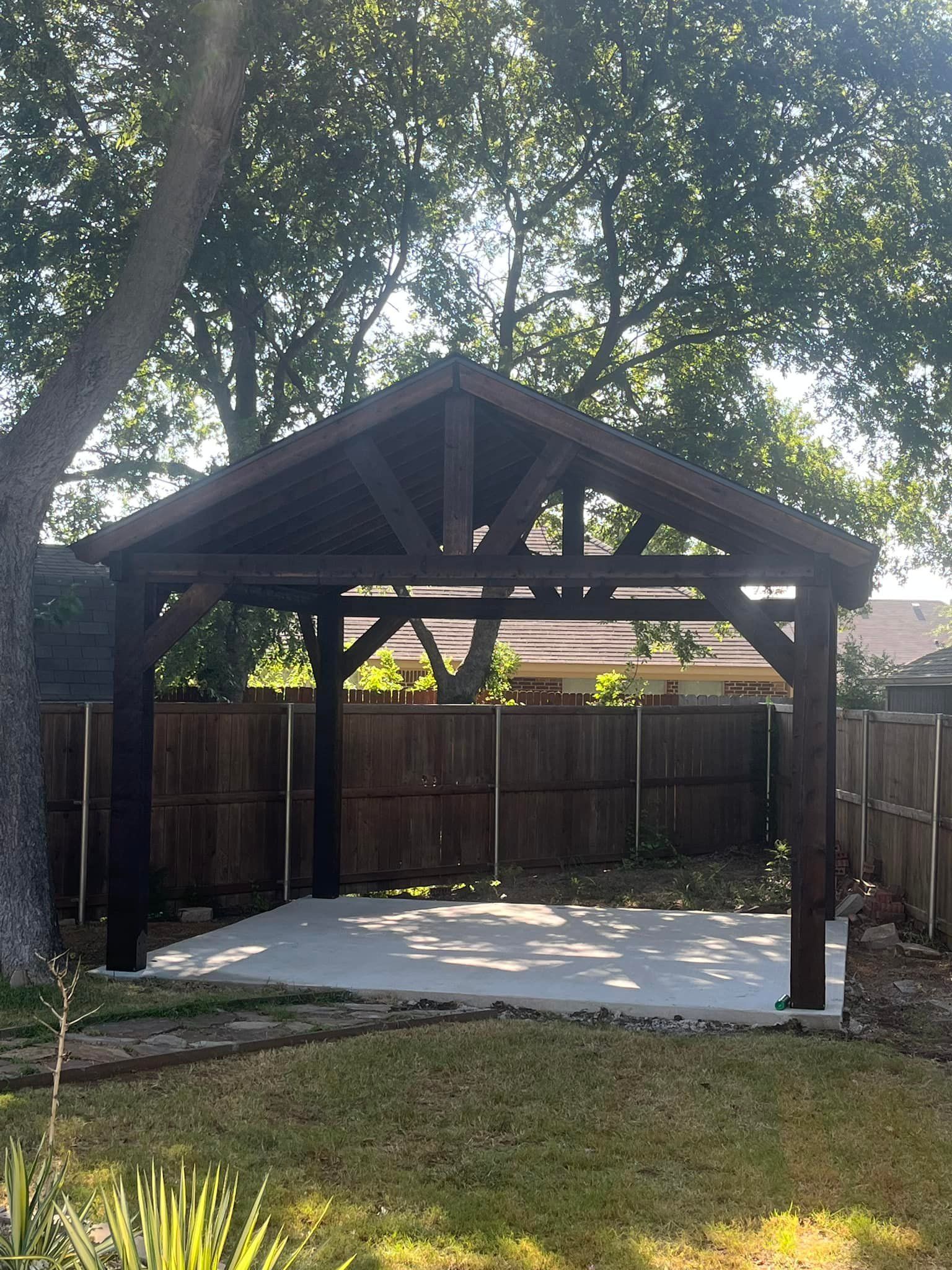 Wooden backyard gazebo with a concrete floor, surrounded by a fence and trees in shade.