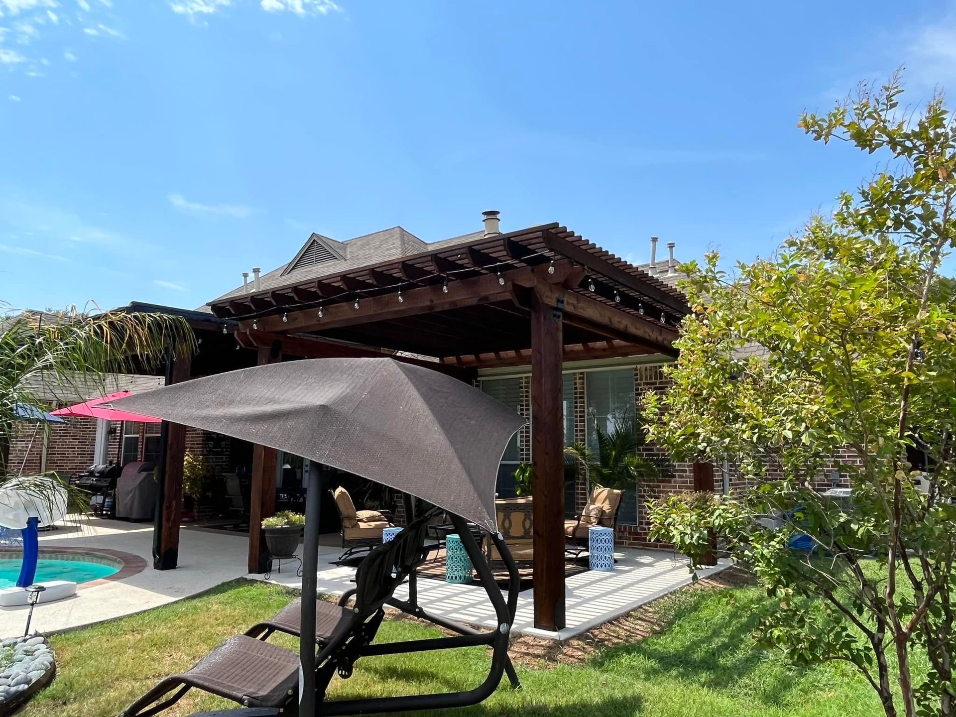 Backyard patio with pergola, shade sail, lounge chair, and pool under a blue sky