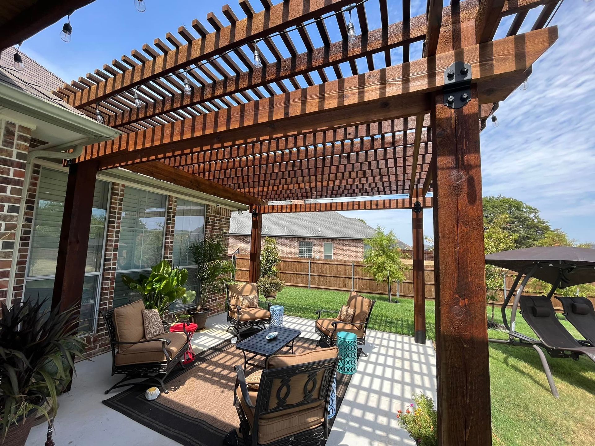 Backyard patio with wooden pergola, cushioned seating, and a grassy lawn under a sunny sky