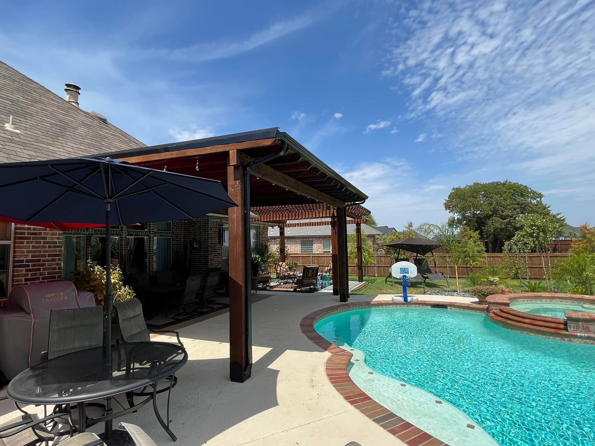 Backyard pool with curved turquoise water, brick deck, and shaded patio seating under a blue sky