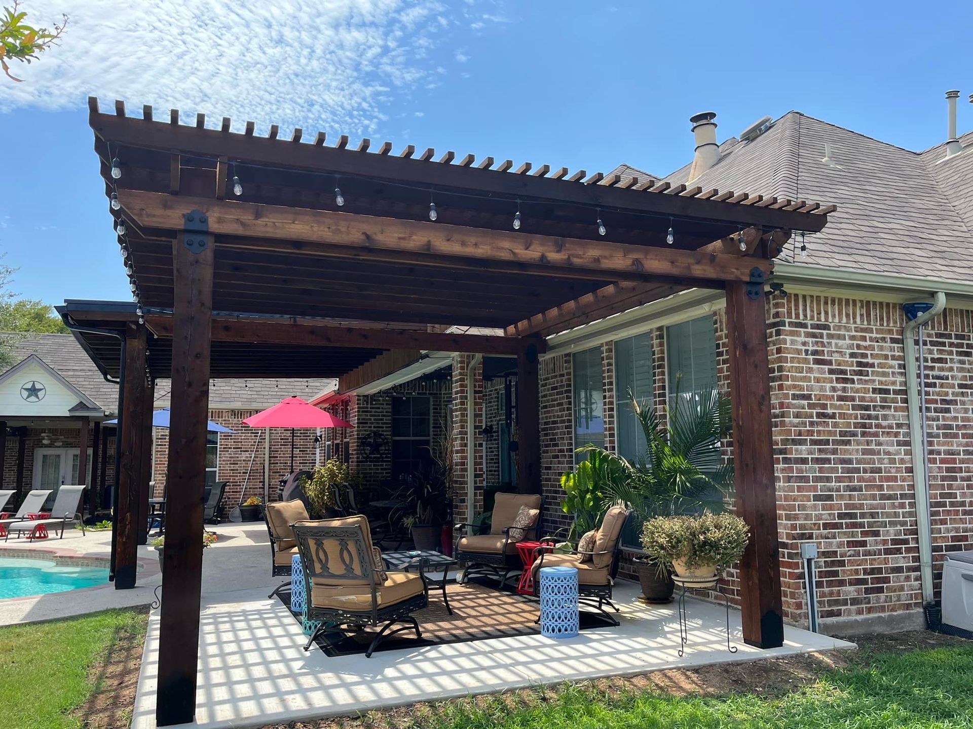 Outdoor patio with wooden pergola, cushioned seating, red umbrella, and brick house wall under a blue sky