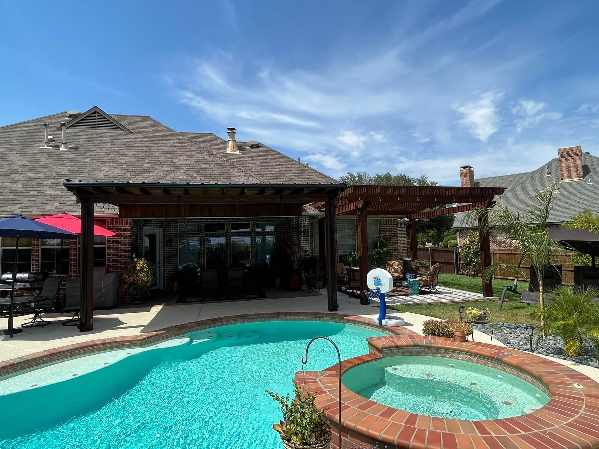 Backyard pool and hot tub beside a shaded patio and tile-roofed house under a blue sky