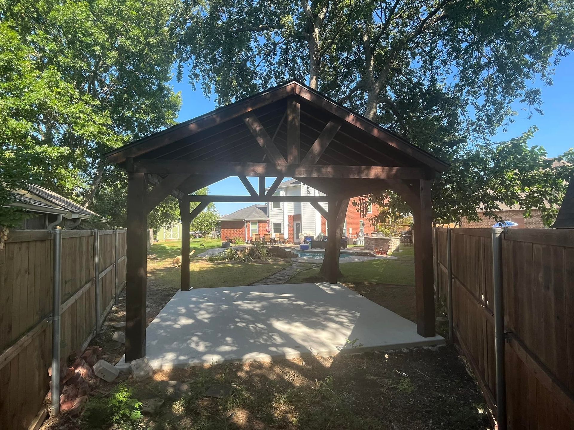 Wooden carport at the end of a fenced backyard path, with a concrete pad and trees overhead.