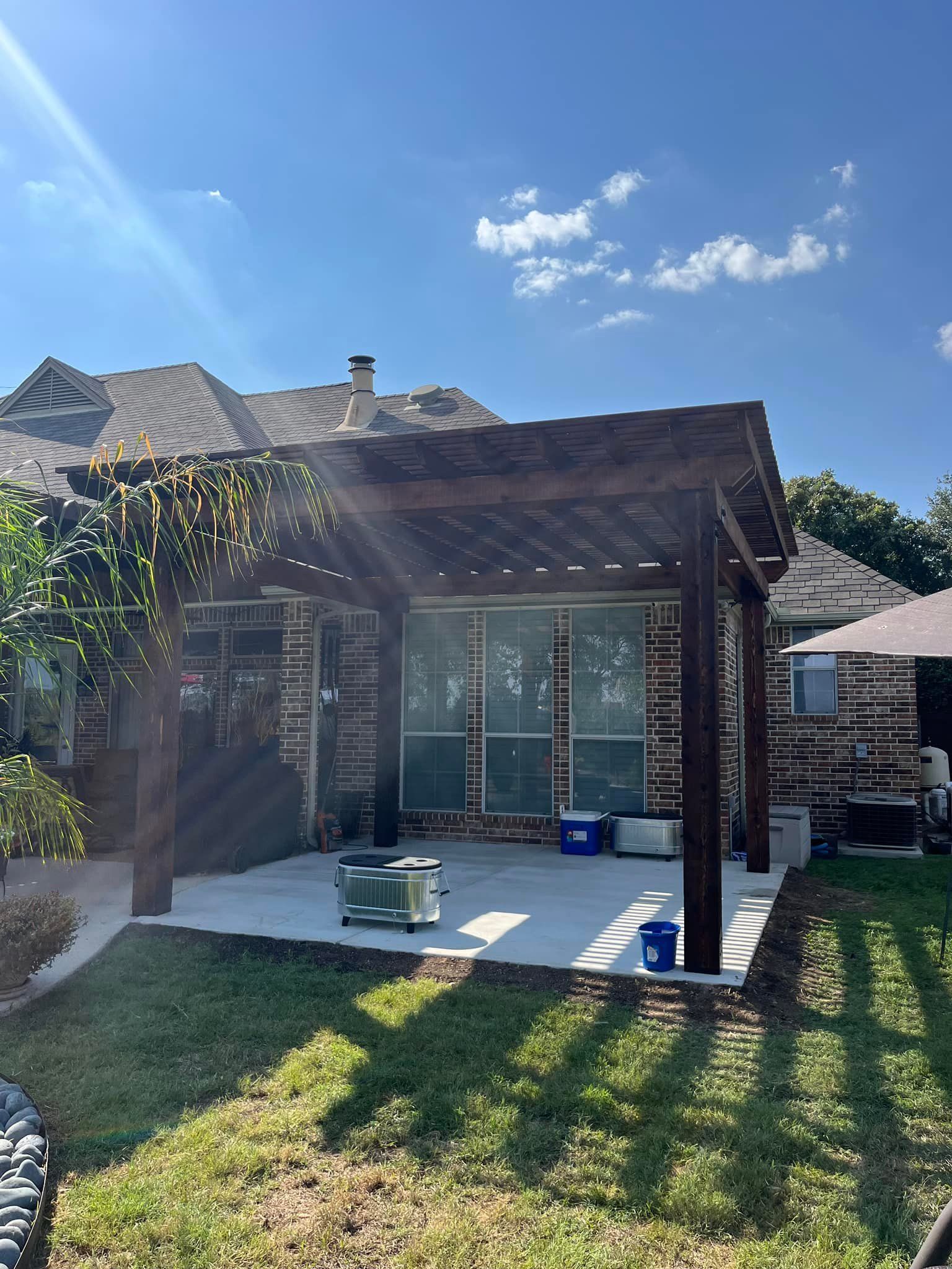 Small stone patio with a pergola, table and chairs, beside a lawn under a bright blue sky