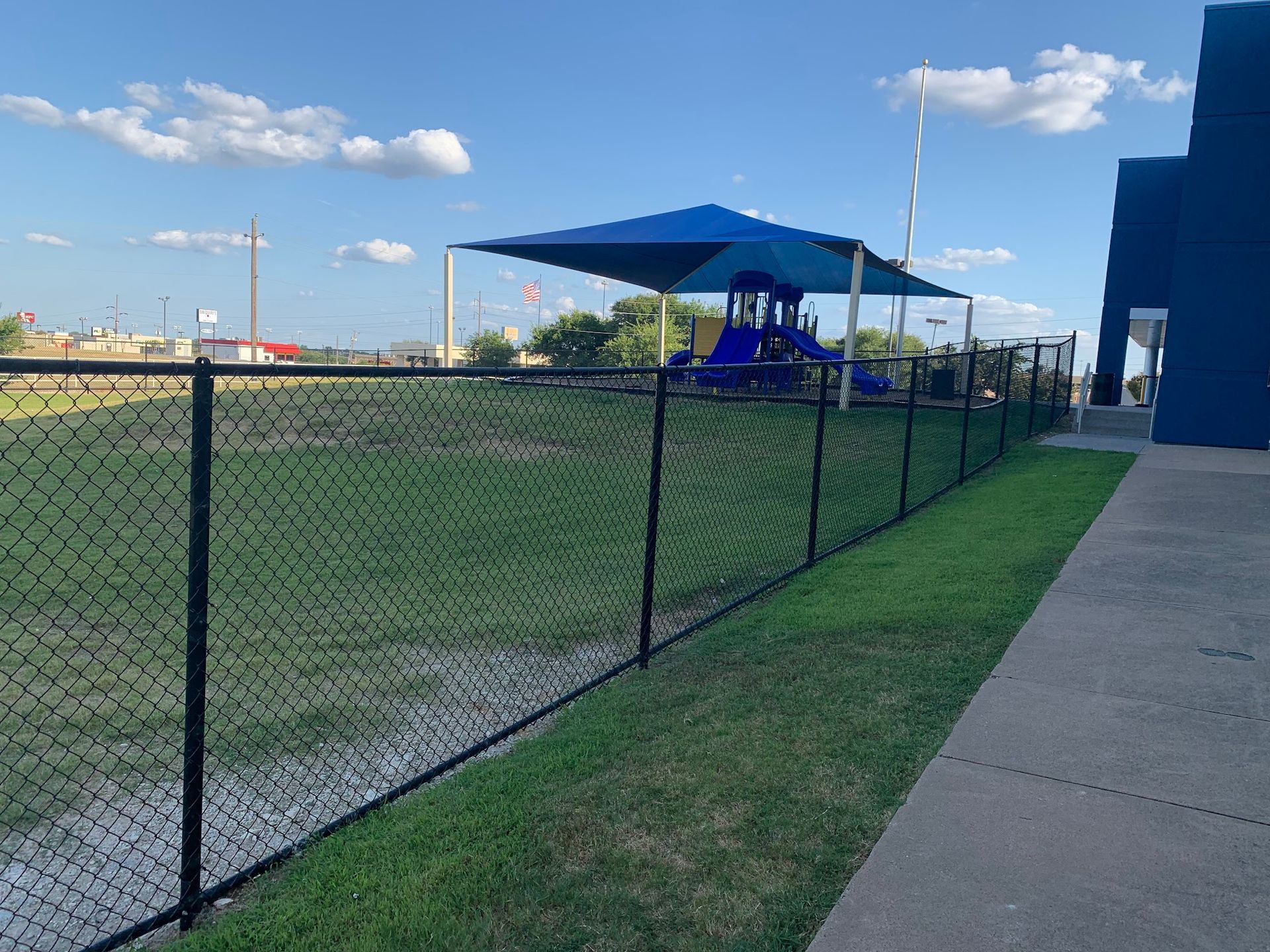 Chain-link fence beside a grassy field and sidewalk, with a blue canopy shelter under a sunny sky.