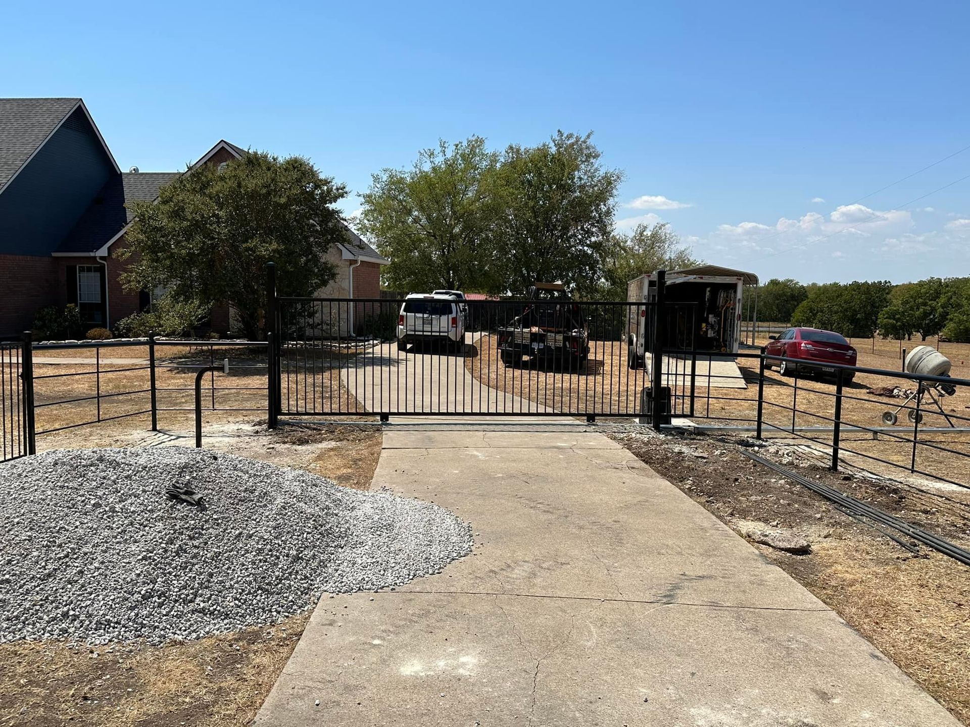 Concrete path to a gated driveway beside a house, gravel, and parked cars on a sunny day