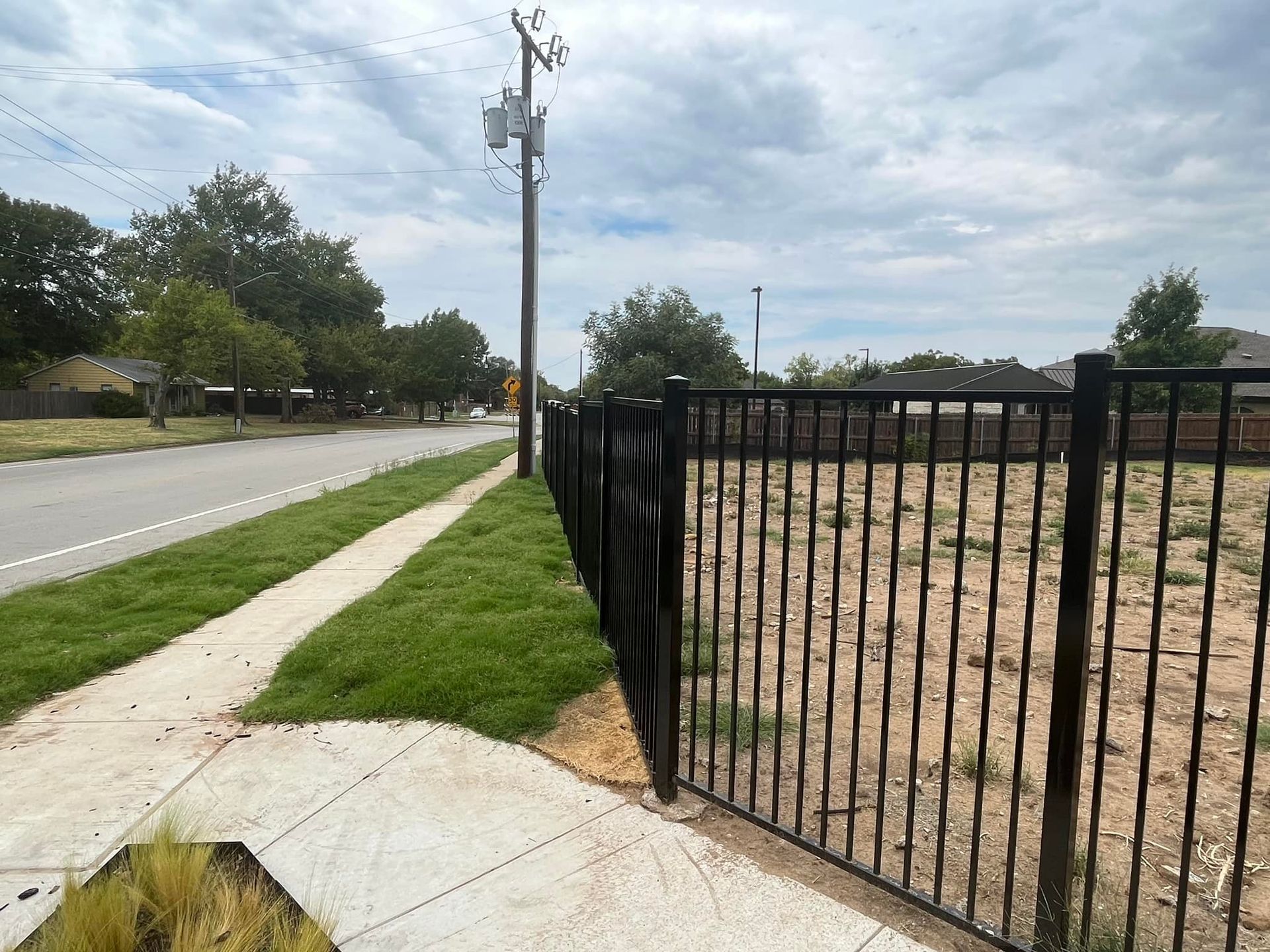 Sidewalk beside a road, black metal fence, and grassy lot under a cloudy sky.