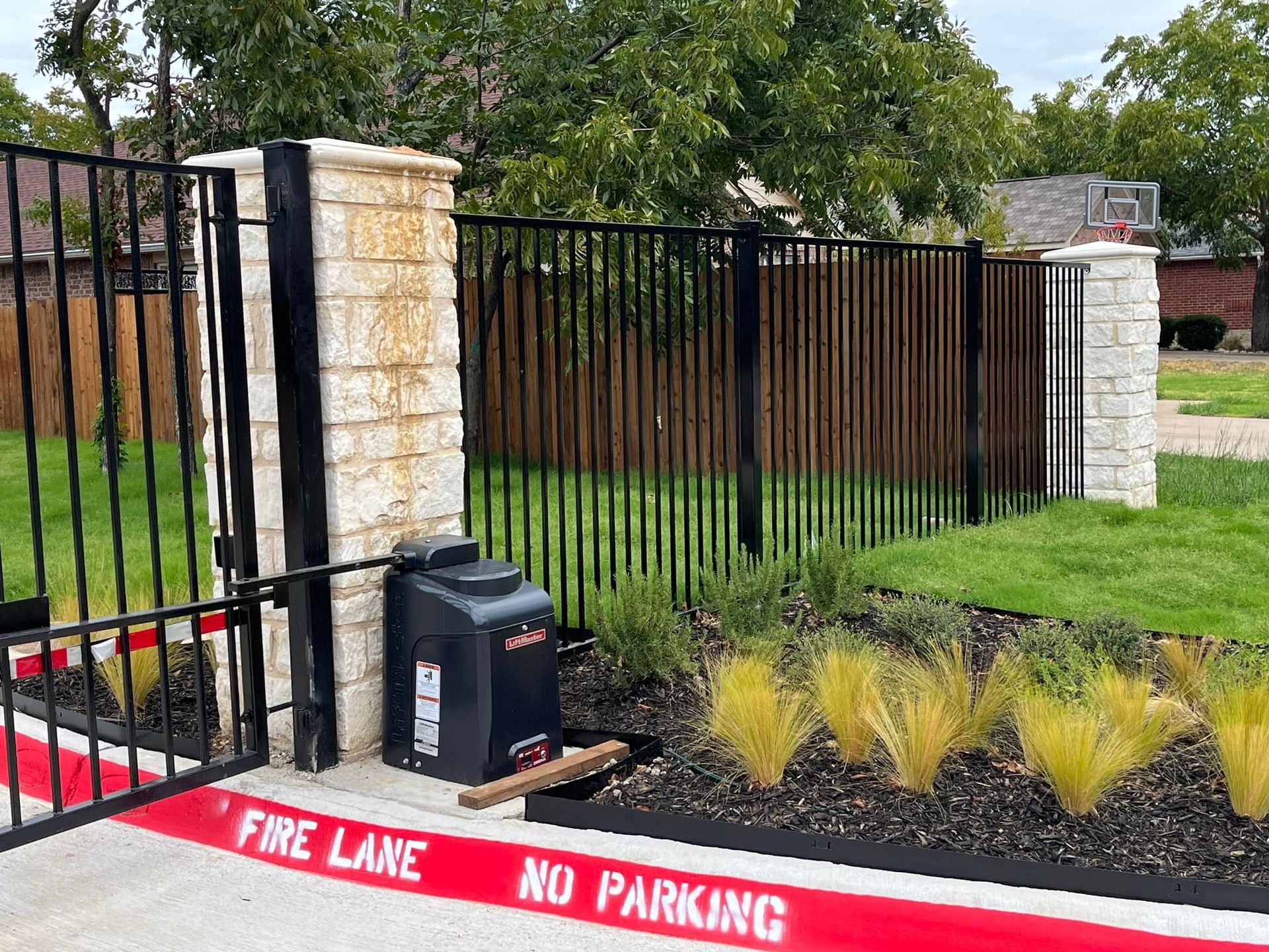 Stone-and-iron fence entrance with red curb reading “FIRE LANE NO PARKING,” trash bin, and mulch bed