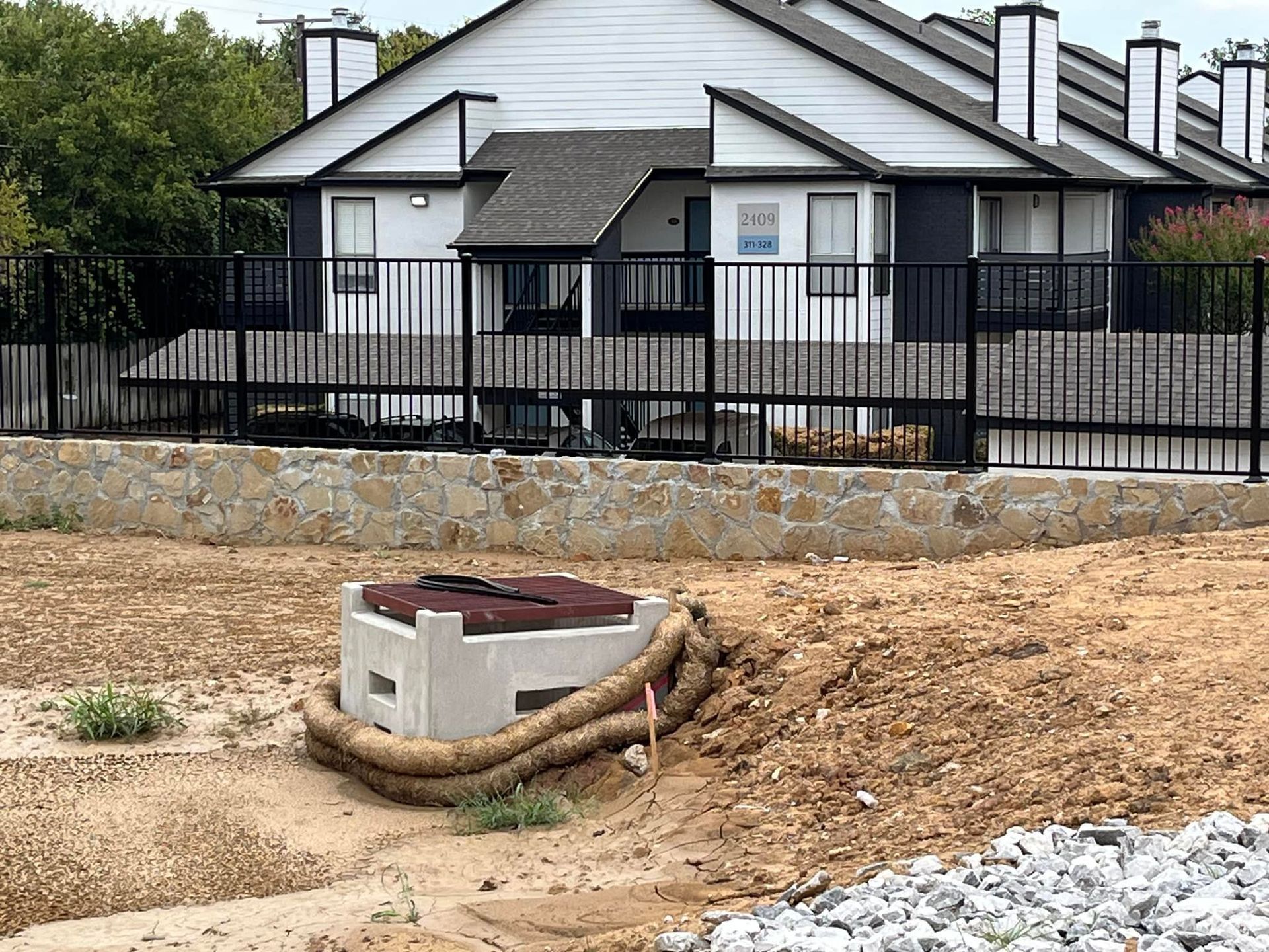 Construction site with a concrete utility box, hose, and a house behind a black fence