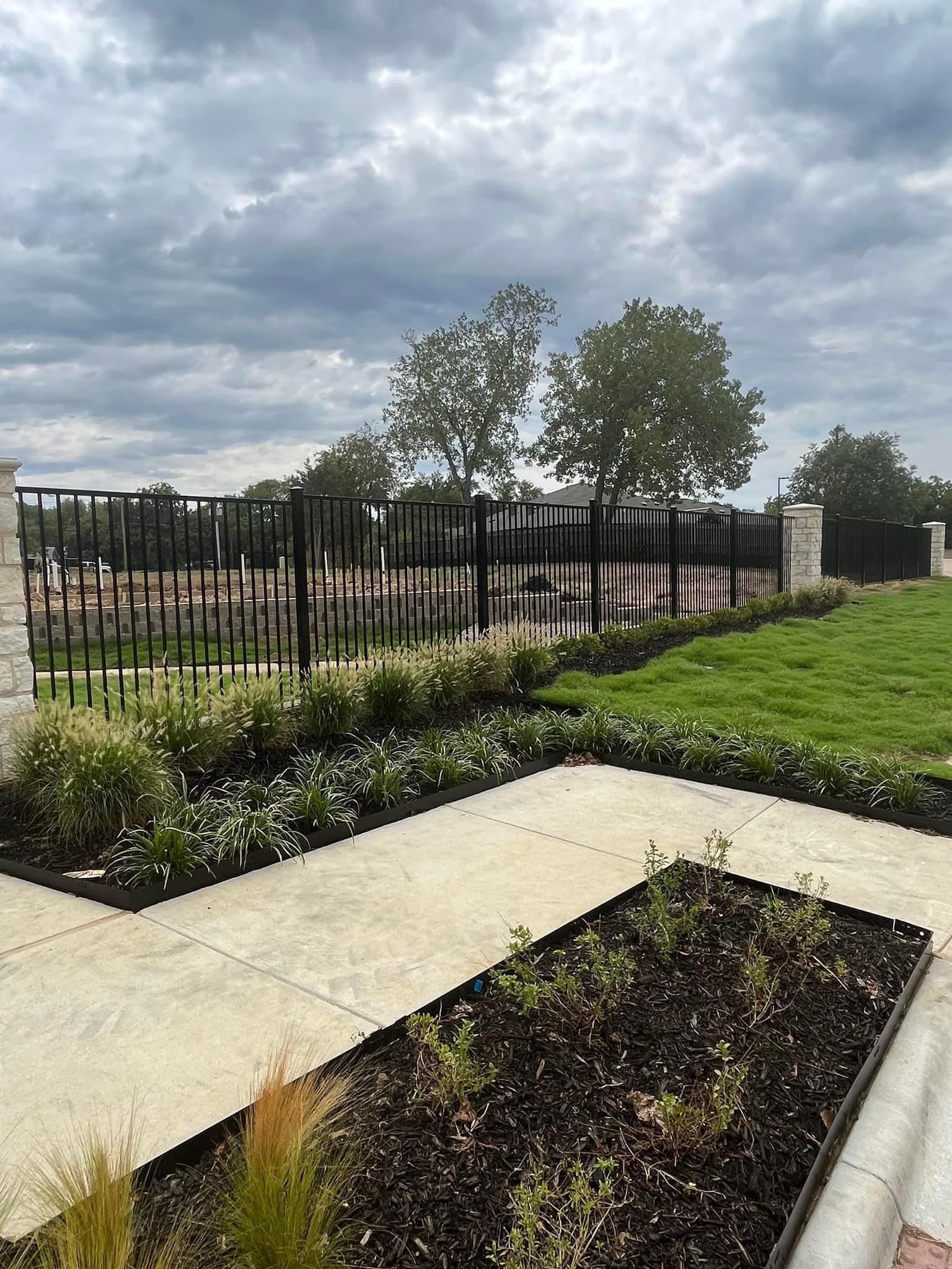 Landscaped garden with concrete paths, flower beds, black fence, and cloudy sky