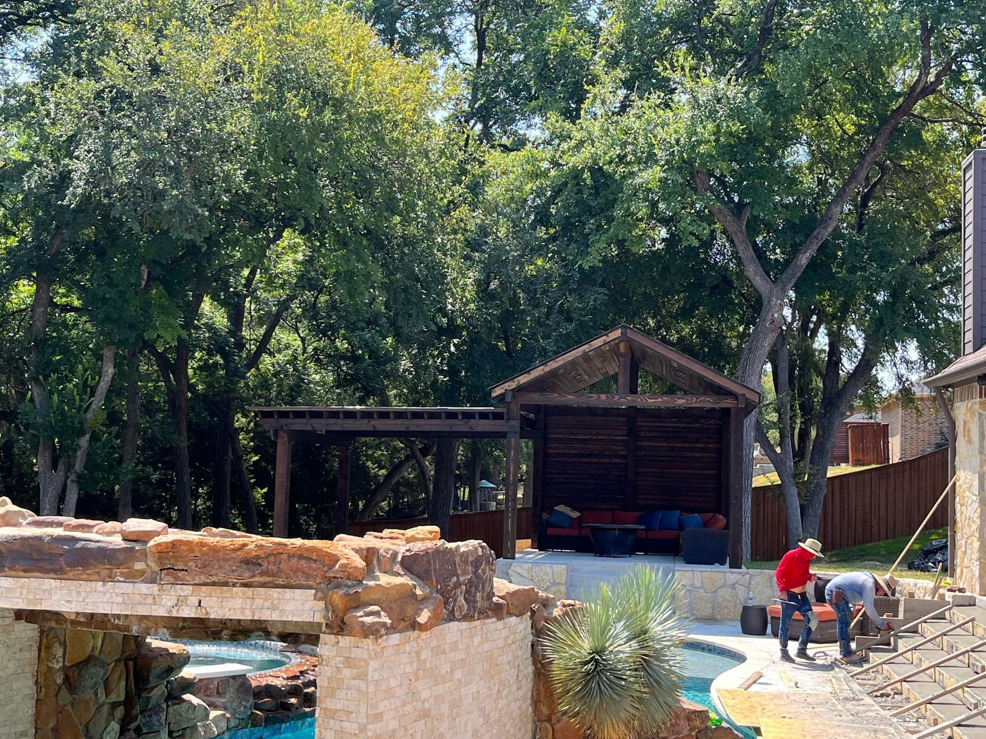 Backyard pool with a stone waterfall, wooden gazebo, and two people working near the water