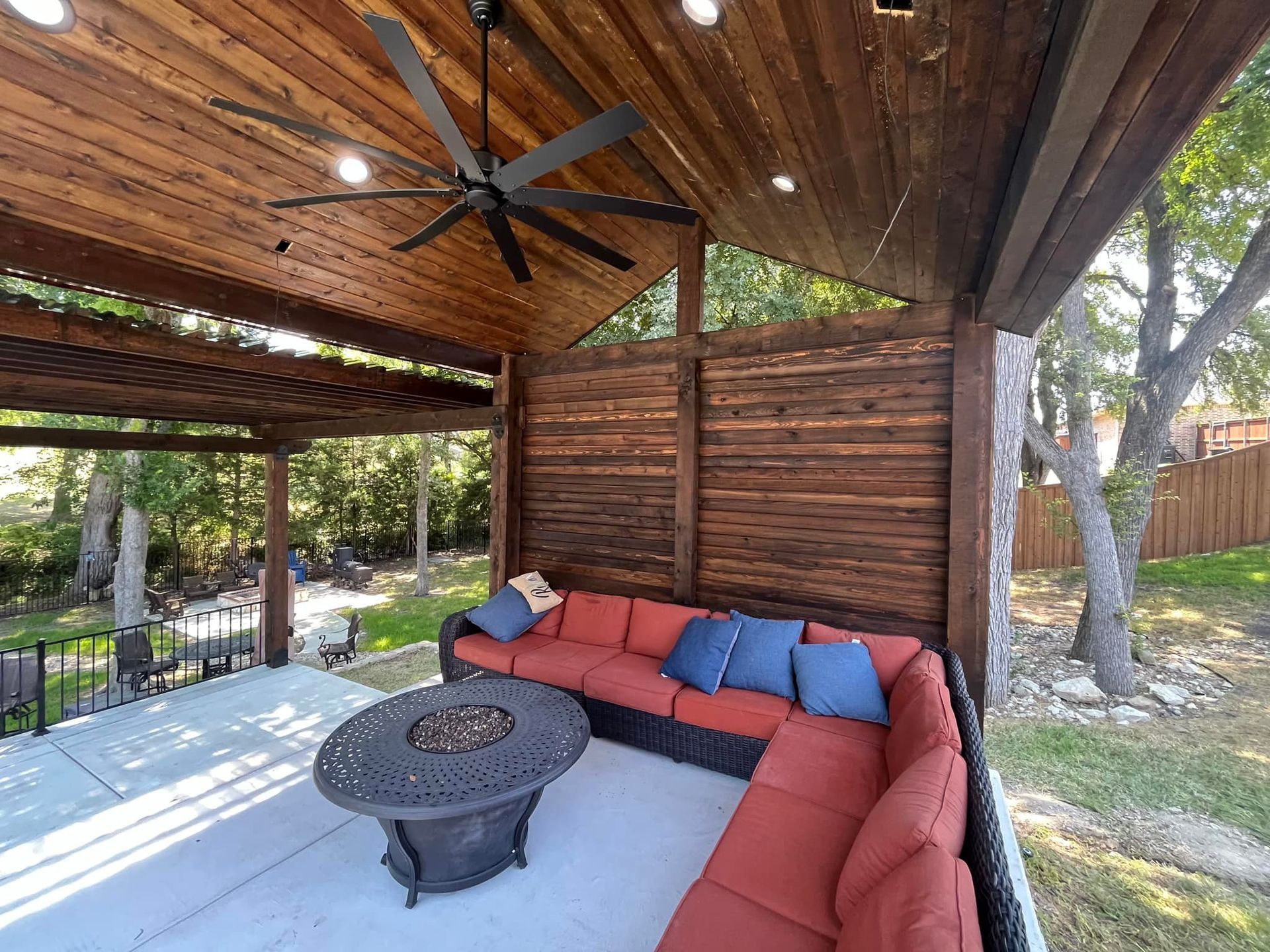 Covered patio with red sectional, blue pillows, round fire pit table, wood ceiling, and ceiling fan
