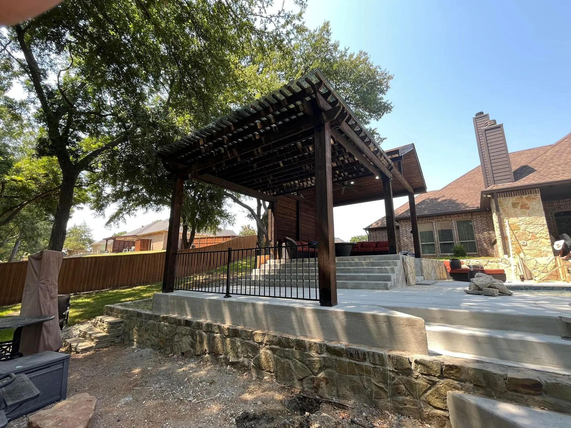 Shaded outdoor pavilion with stone steps, trees, and a brick building under a blue sky
