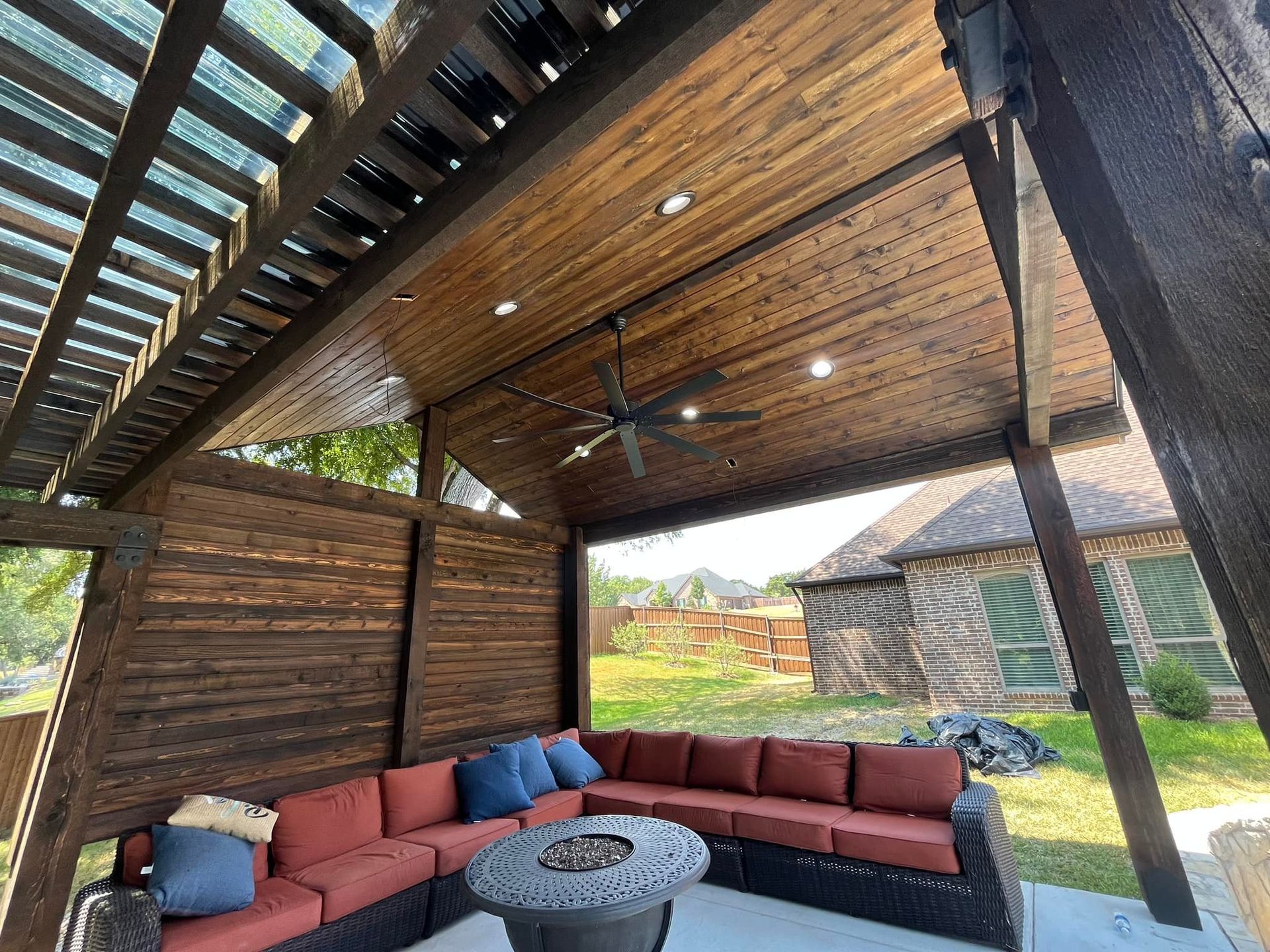 Covered wooden patio with red sectional sofa, round fire pit table, and ceiling fan