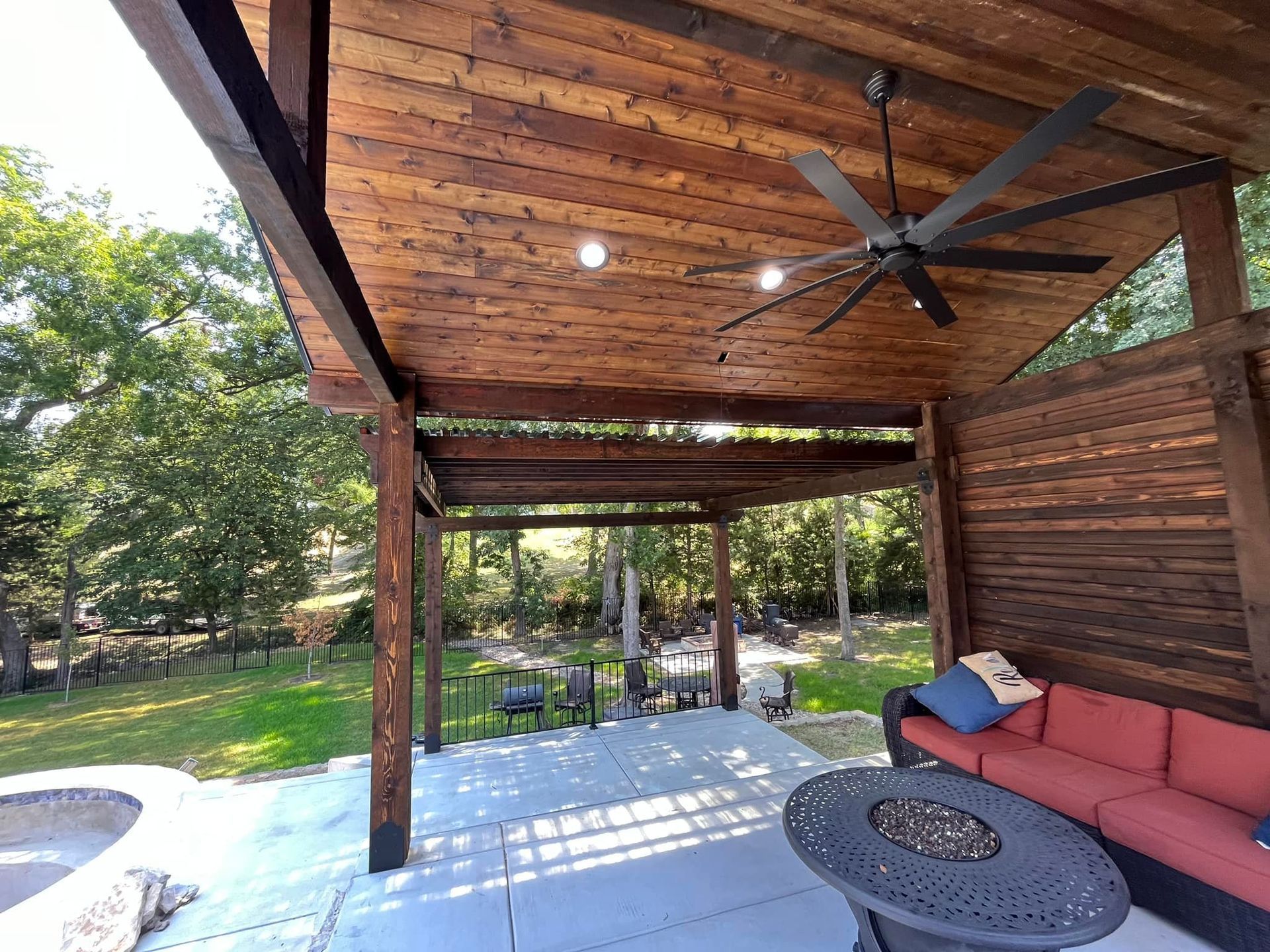 Covered wooden patio with a ceiling fan, red sofa, round fire pit table, and backyard view.