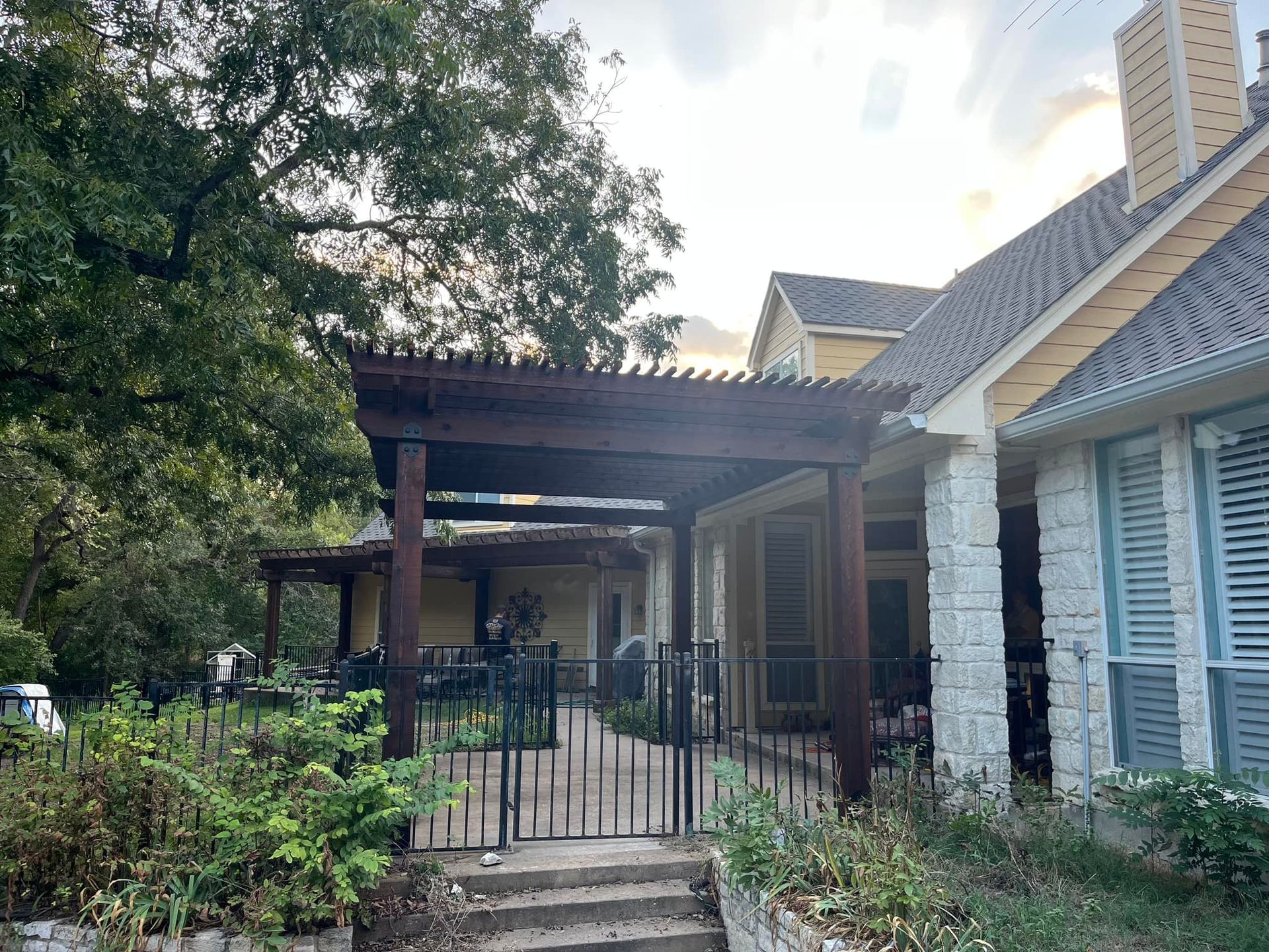 Front porch with a black metal gate, wooden pergola, and blue-shuttered house beside leafy trees