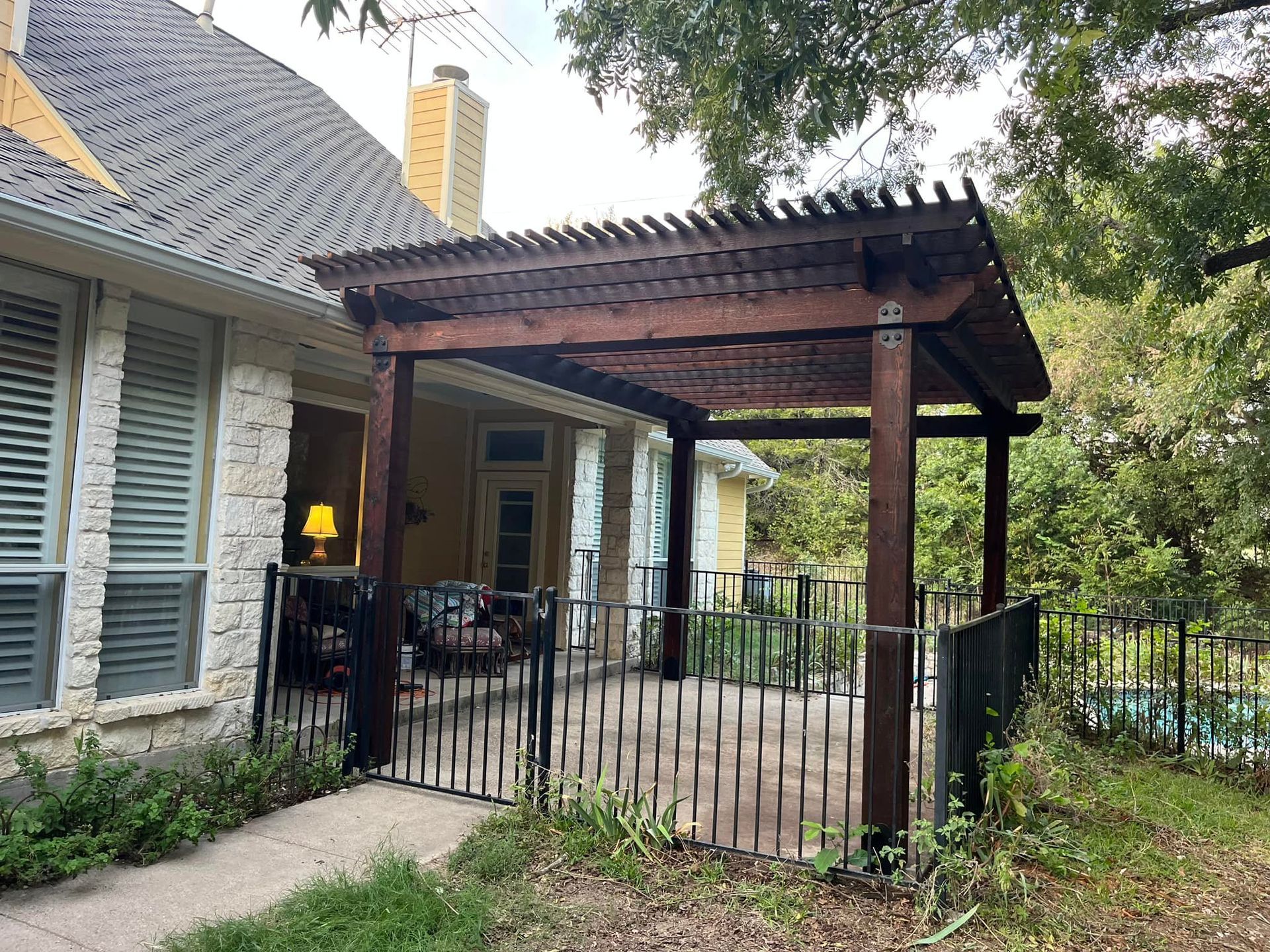 Covered patio with wooden pergola, black railing, and stone house exterior surrounded by greenery