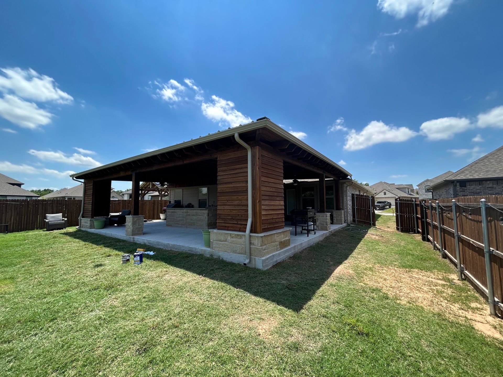 Backyard patio with a brick and wood pavilion under a blue sky, surrounded by grass and fences