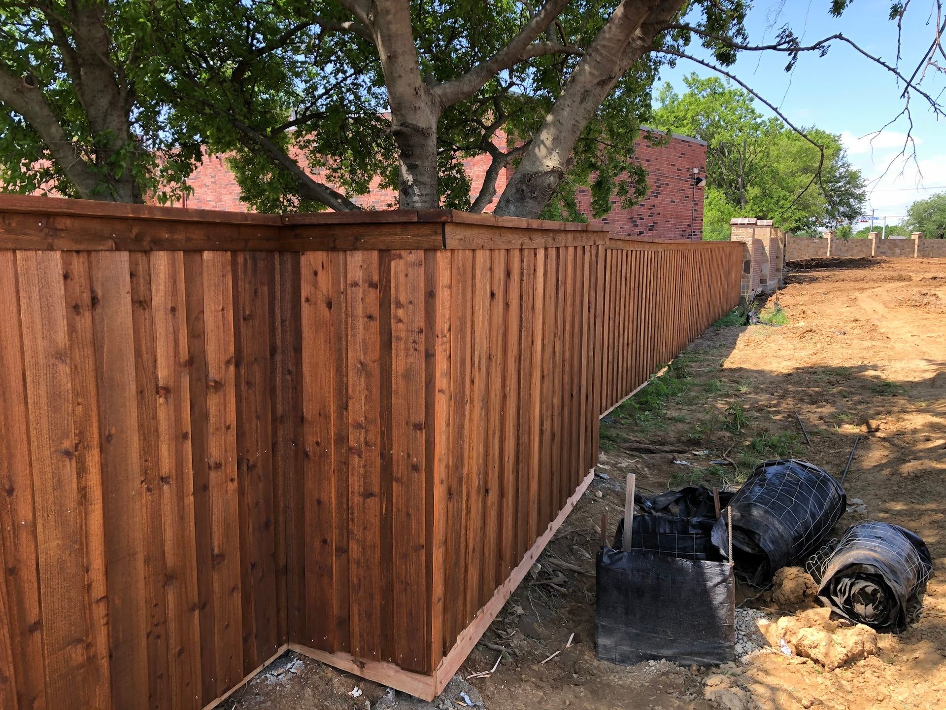 New wooden privacy fence along a dirt path, with trees and black trash bags nearby
