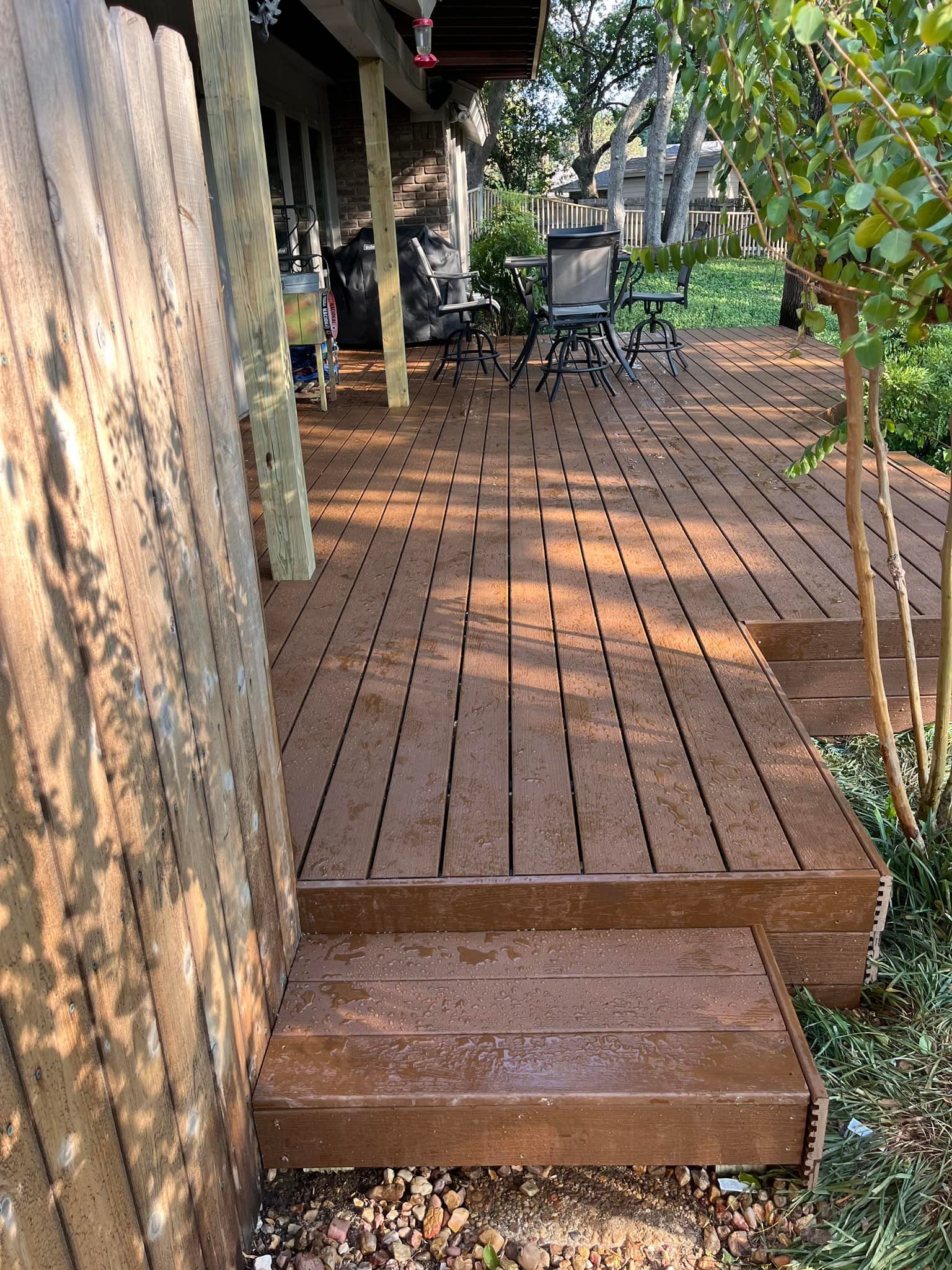 Wooden deck walkway beside a house, with patio chairs and trees in the background.