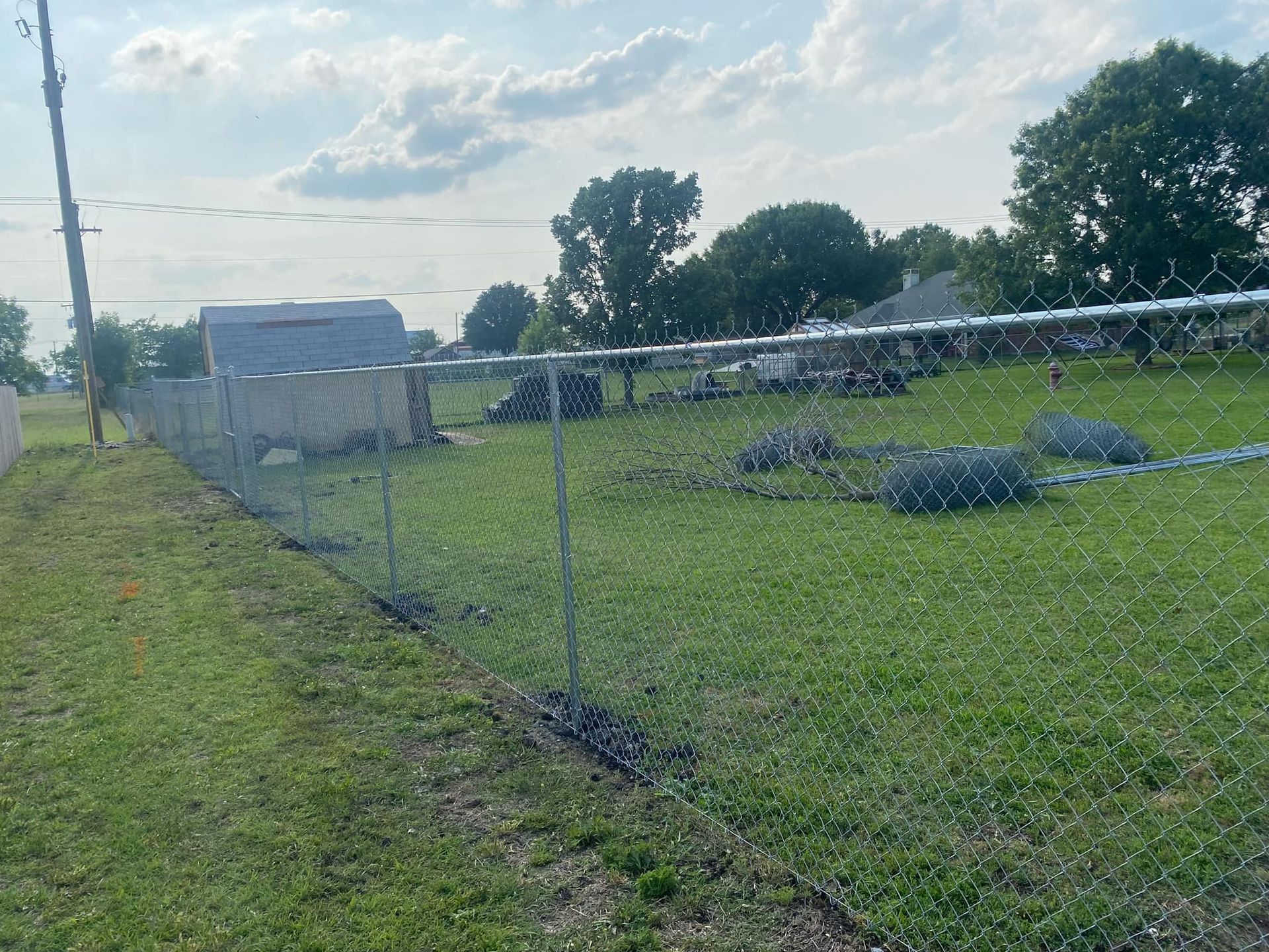 Chain-link fence around a grassy yard with a small barn and trees under a cloudy sky.